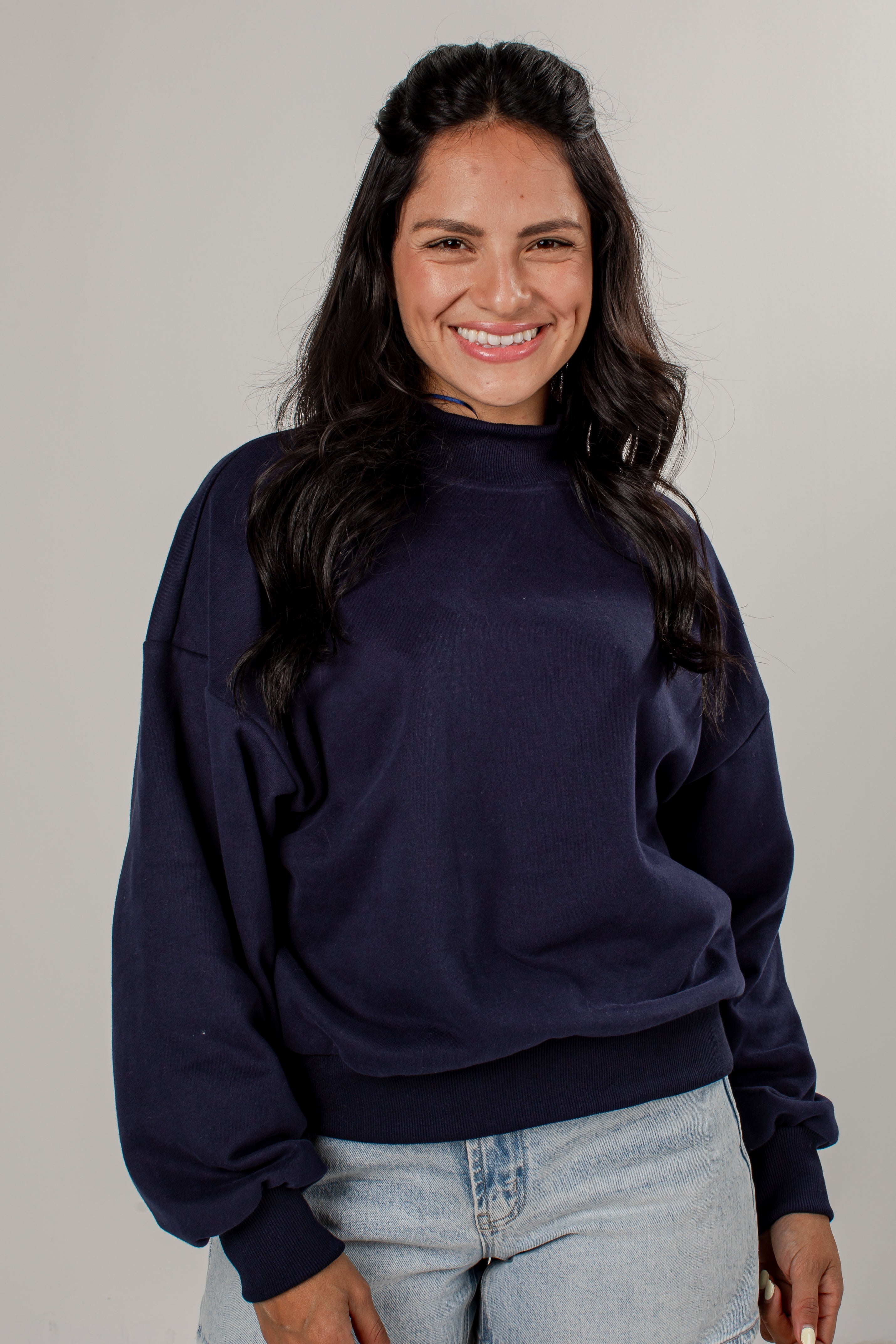A smiling woman with long dark hair wearing a navy blue mock neck sweatshirt and light wash jeans, standing against a plain light gray studio background.