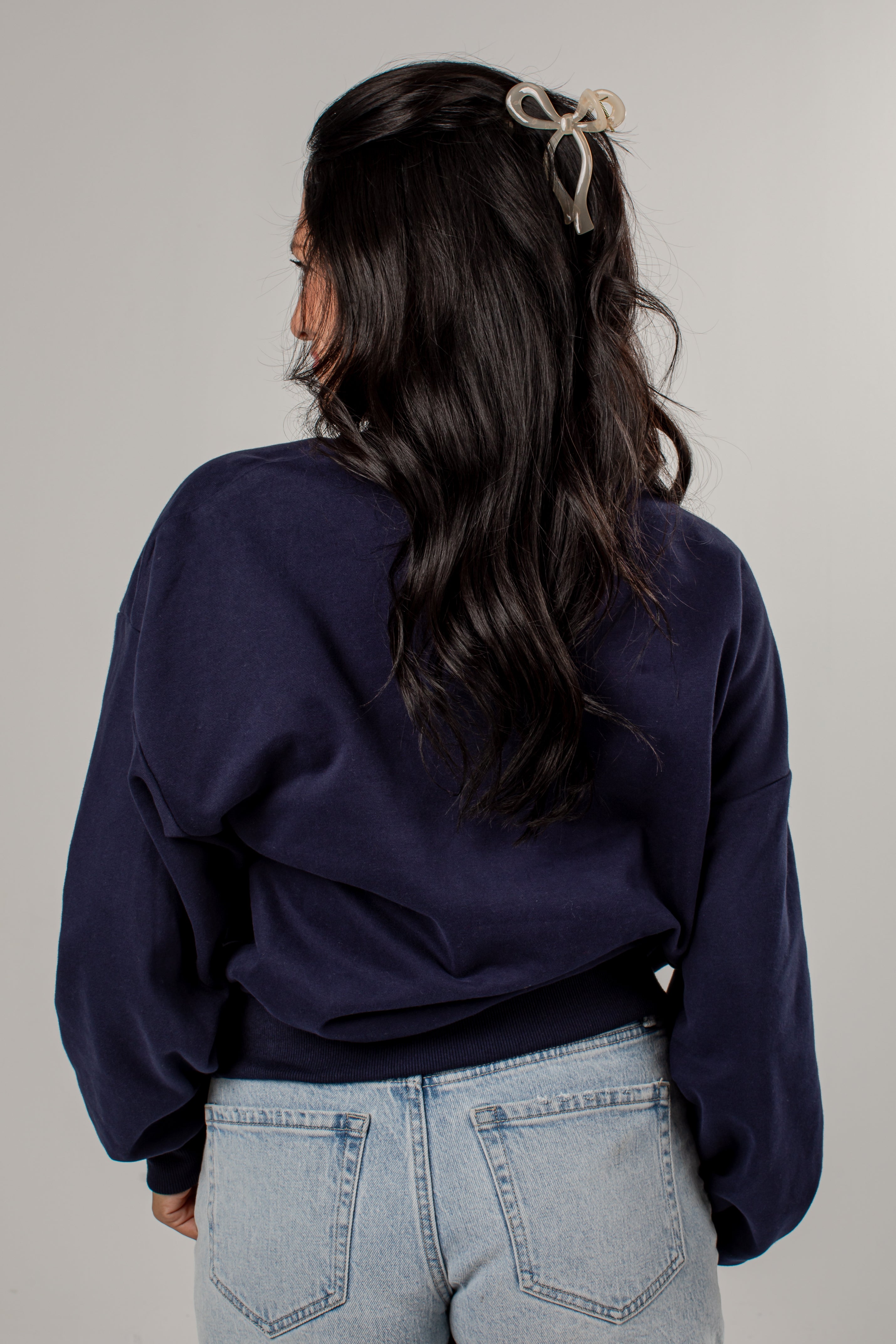 A woman shown from behind wearing a dark navy blue cropped sweatshirt and light wash blue jeans, with a beige bow hair clip in her hair, against a plain light gray background.