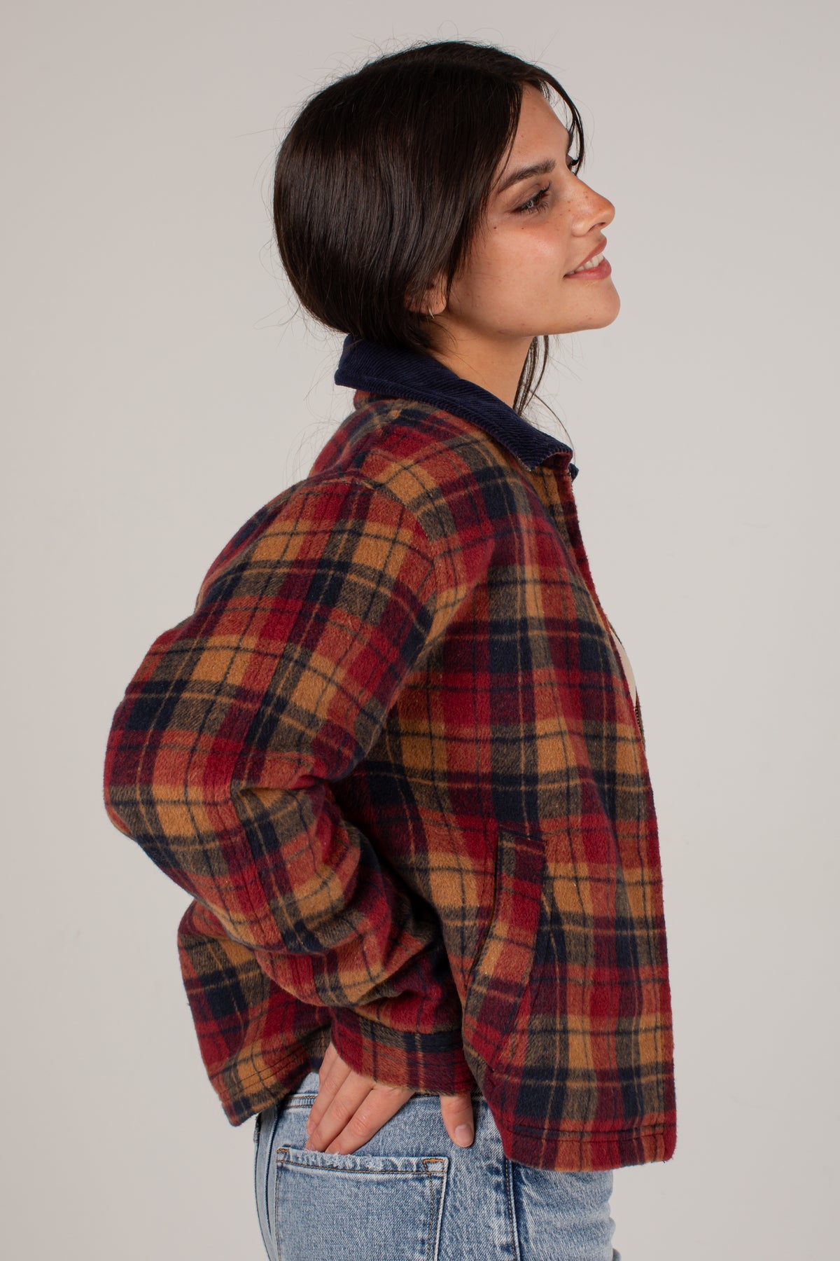 A woman shown in profile wearing a red, navy, and tan plaid flannel shirt jacket over a dark navy collared shirt, paired with light blue jeans, against a plain light gray studio background.