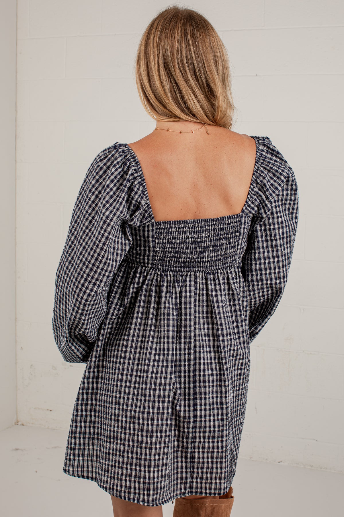 A woman shown from the back wearing a navy and white gingham mini dress with a smocked square neckline and puff sleeves, set against a white paneled wall.