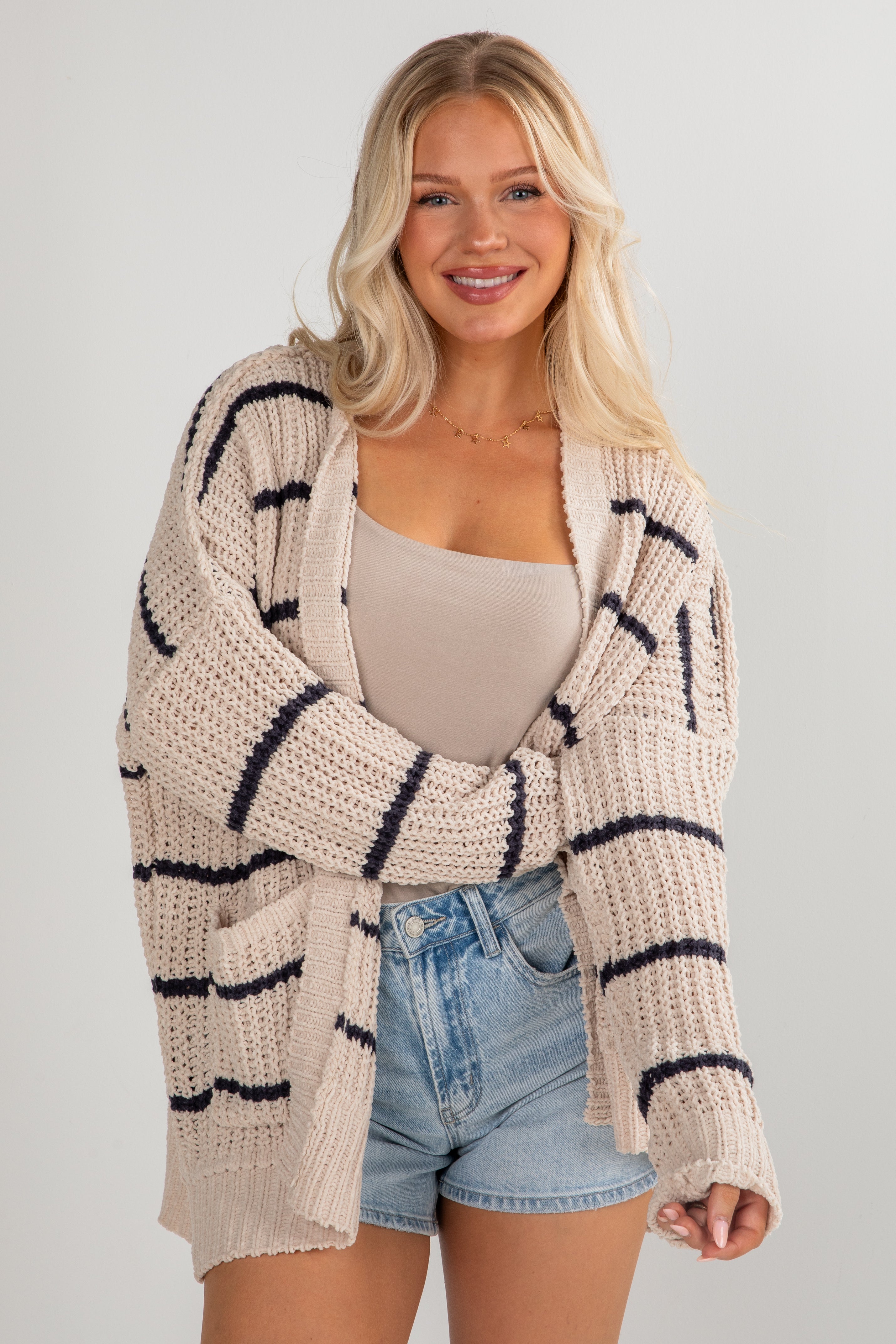 Woman wearing a beige cardigan with black stripes, beige top, and denim shorts on a light gray background