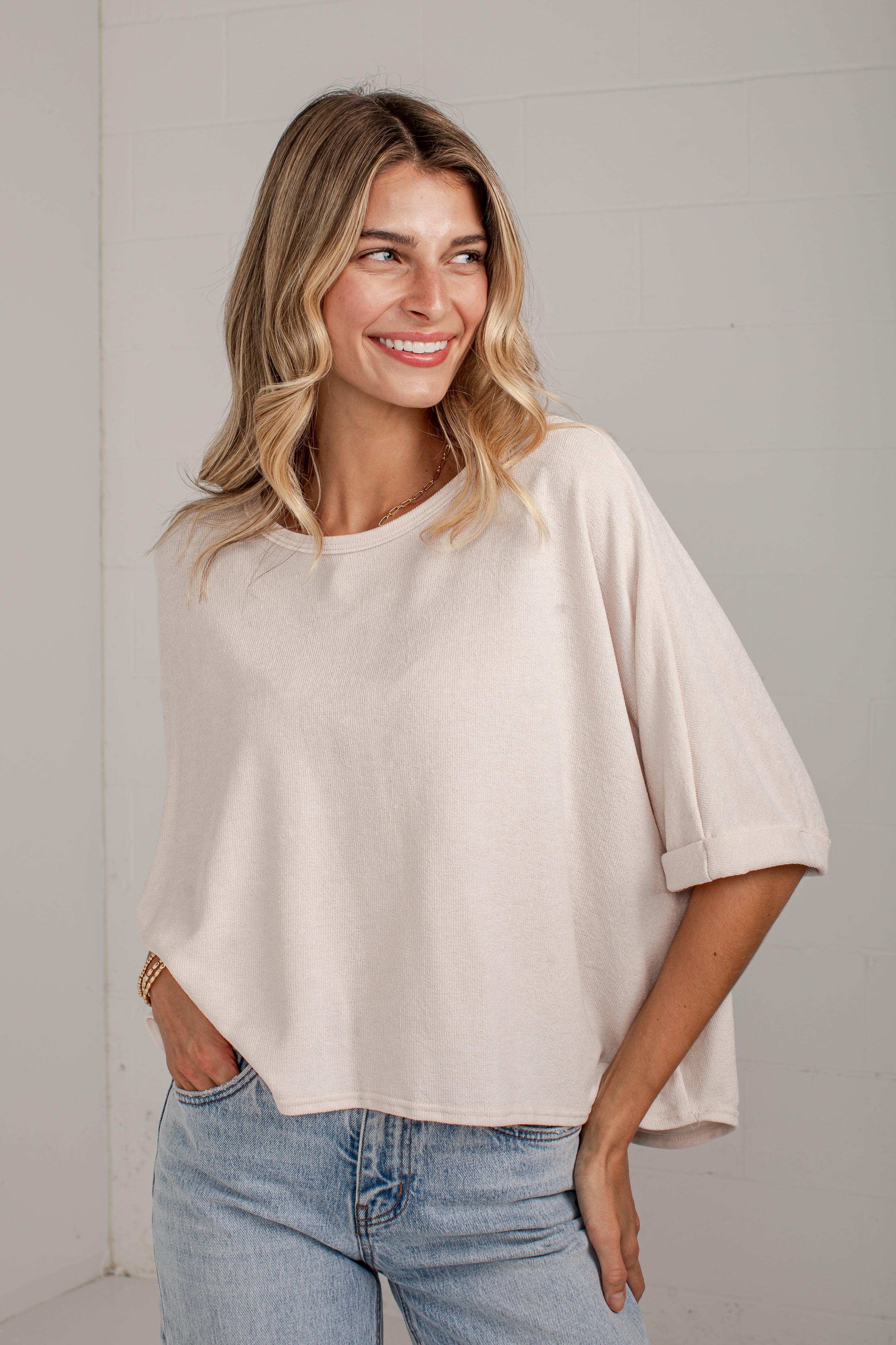 A woman wearing a light beige, oversized short-sleeve top with rolled cuffs and light wash blue jeans, standing against a light gray wall.
