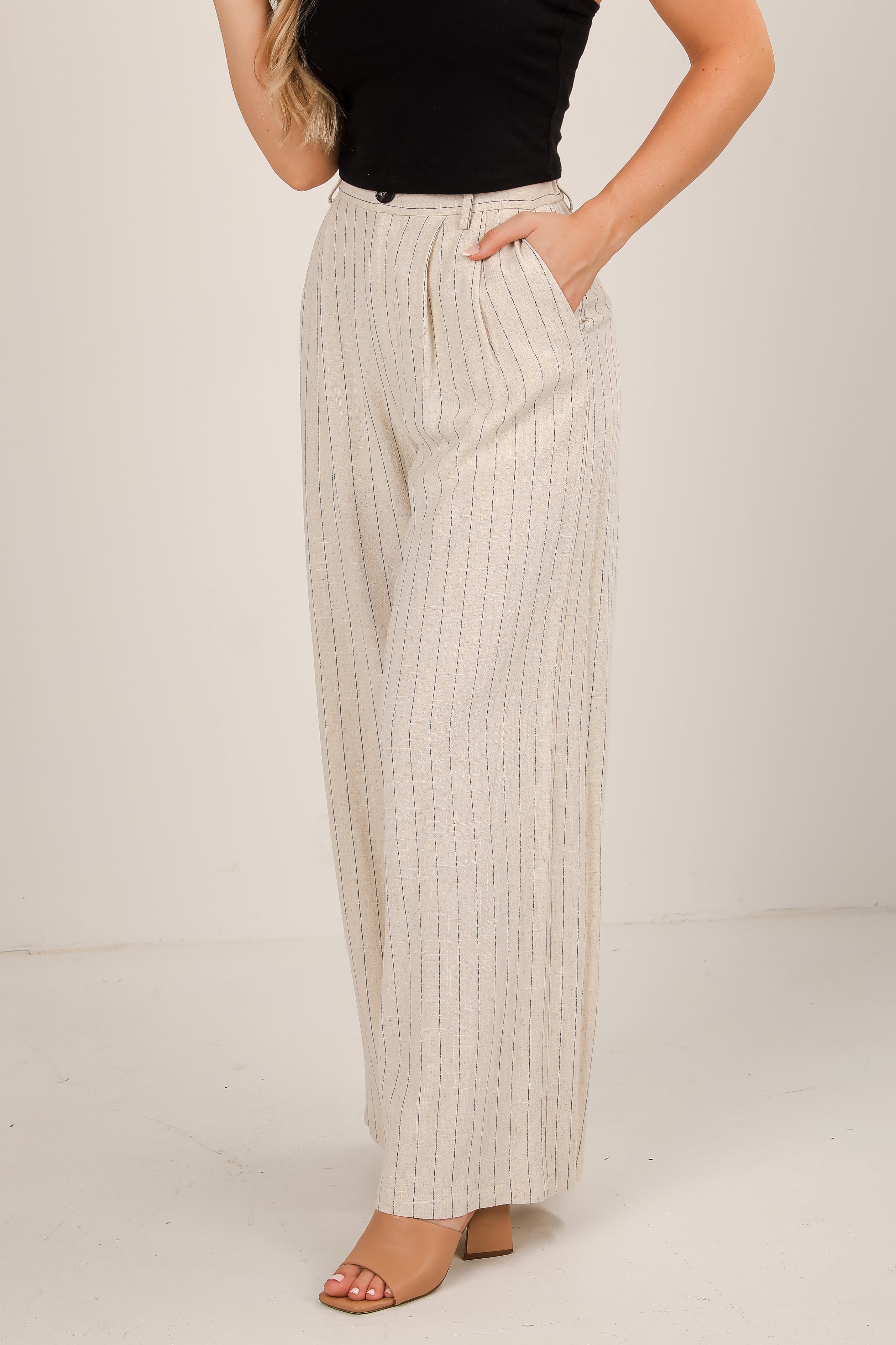 Woman wearing beige pleated pants and a black top on a plain background