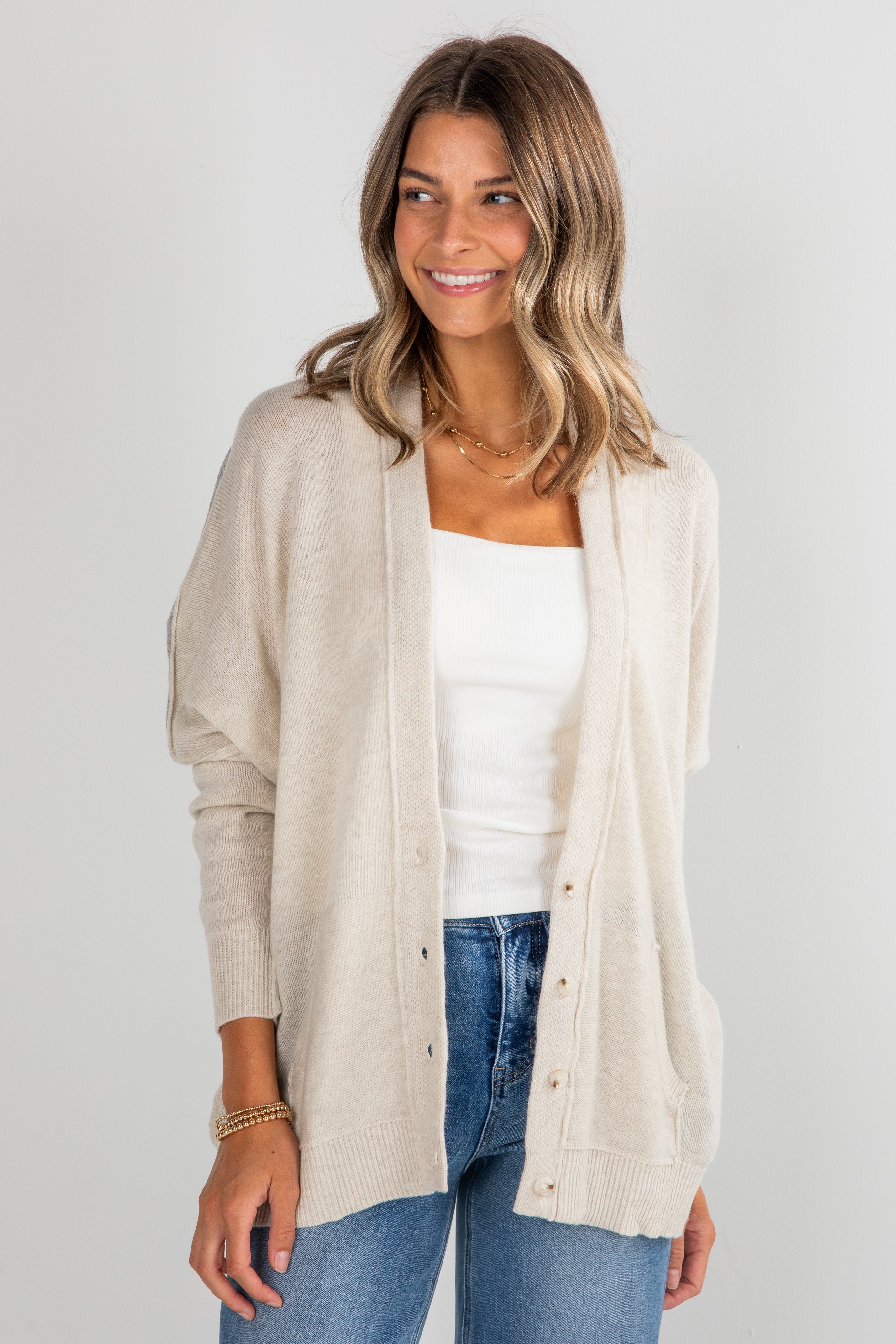 Woman wearing a beige cardigan over a white top and blue jeans on a plain background