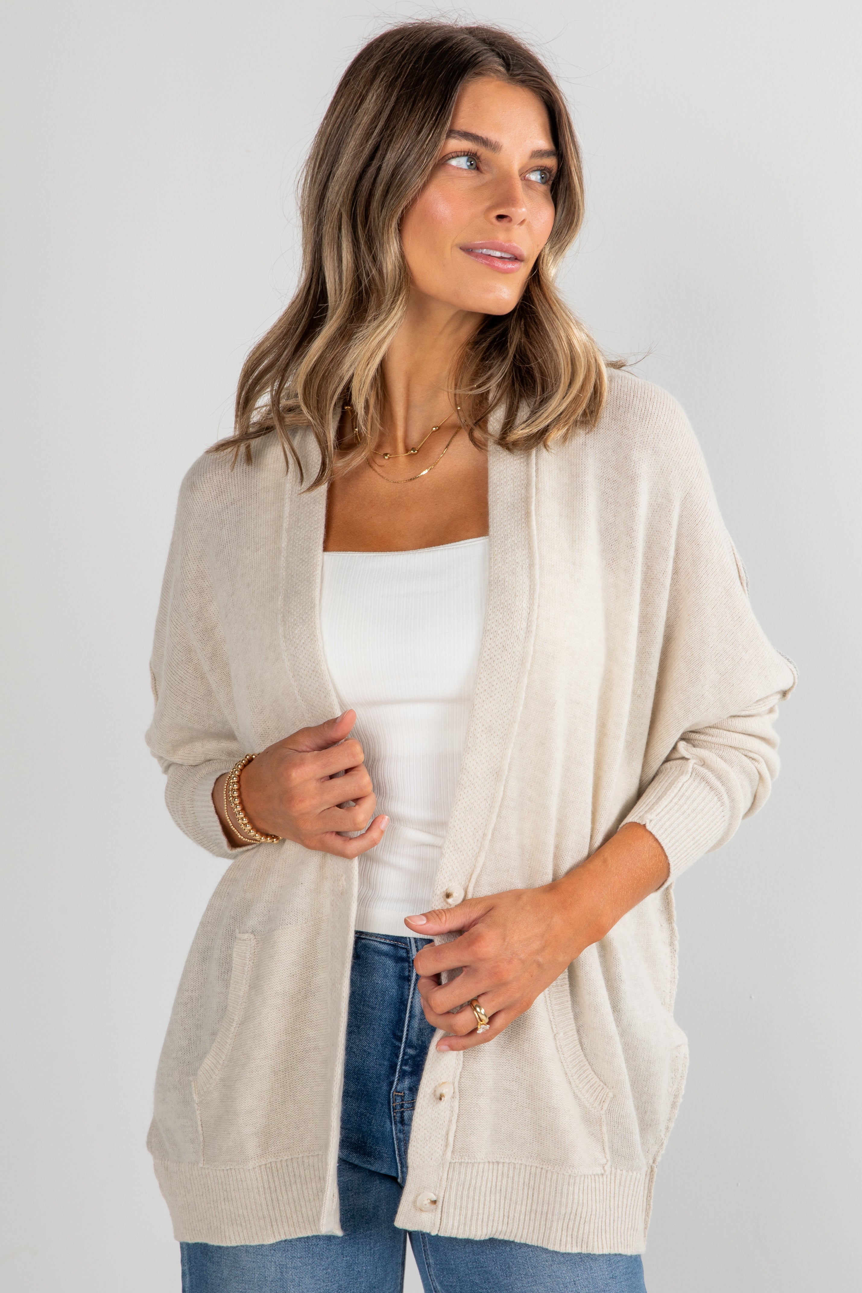 Woman wearing a beige cardigan over a white top and blue jeans on a light gray background