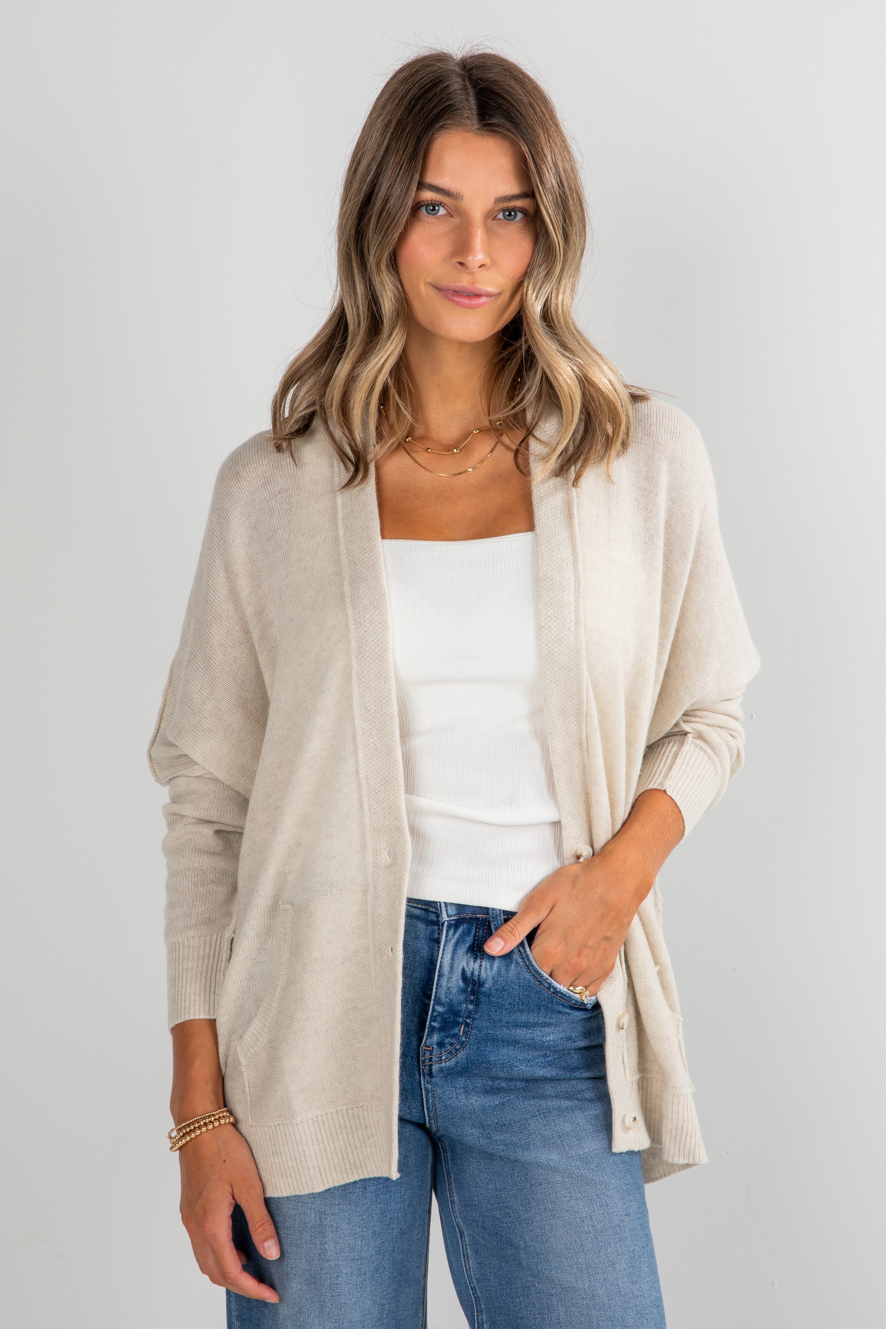 Woman wearing a beige cardigan, white top, and blue jeans on a plain background