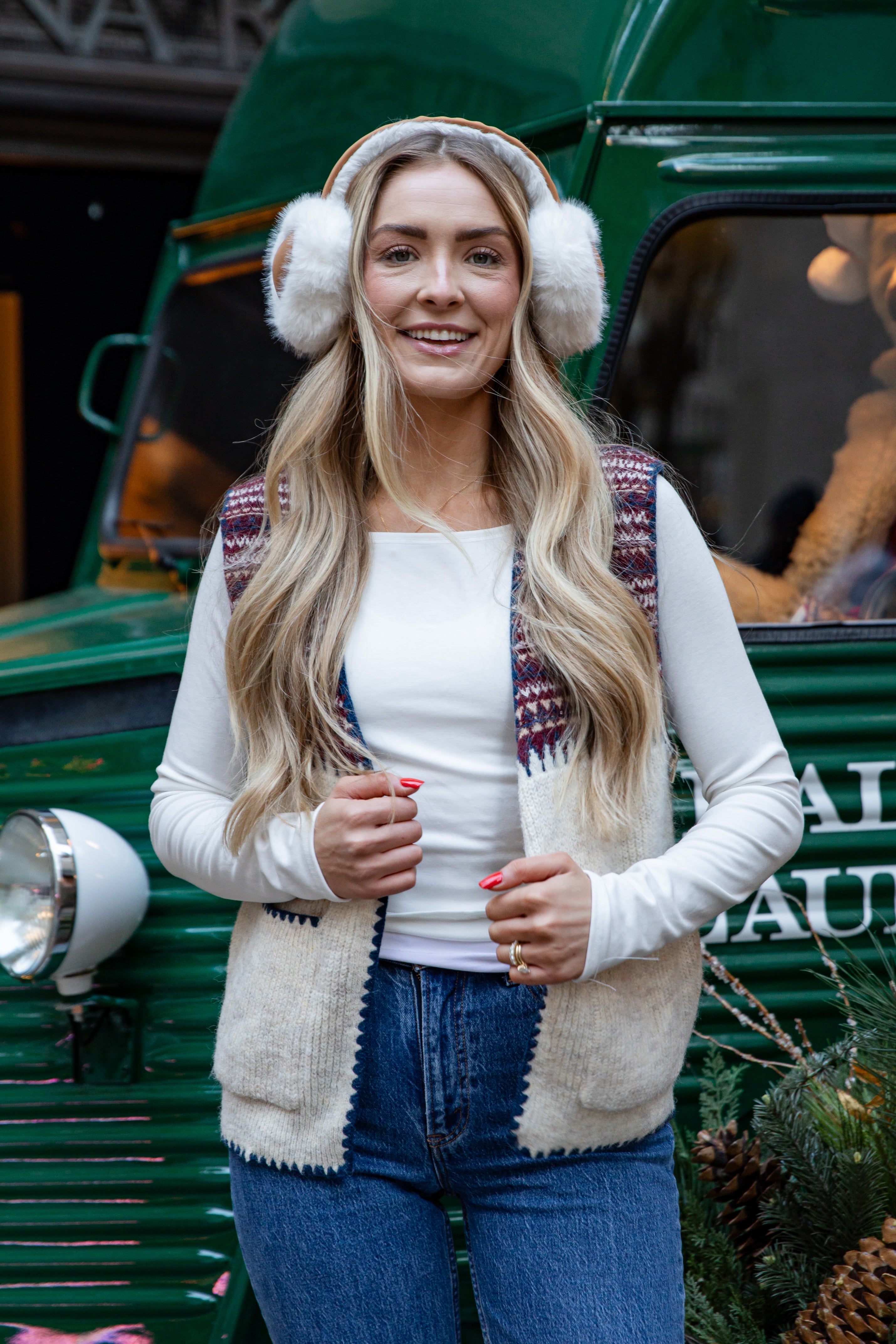 Woman wearing a patterned cardigan, white shirt, blue jeans, and white earmuffs in front of a green vehicle.