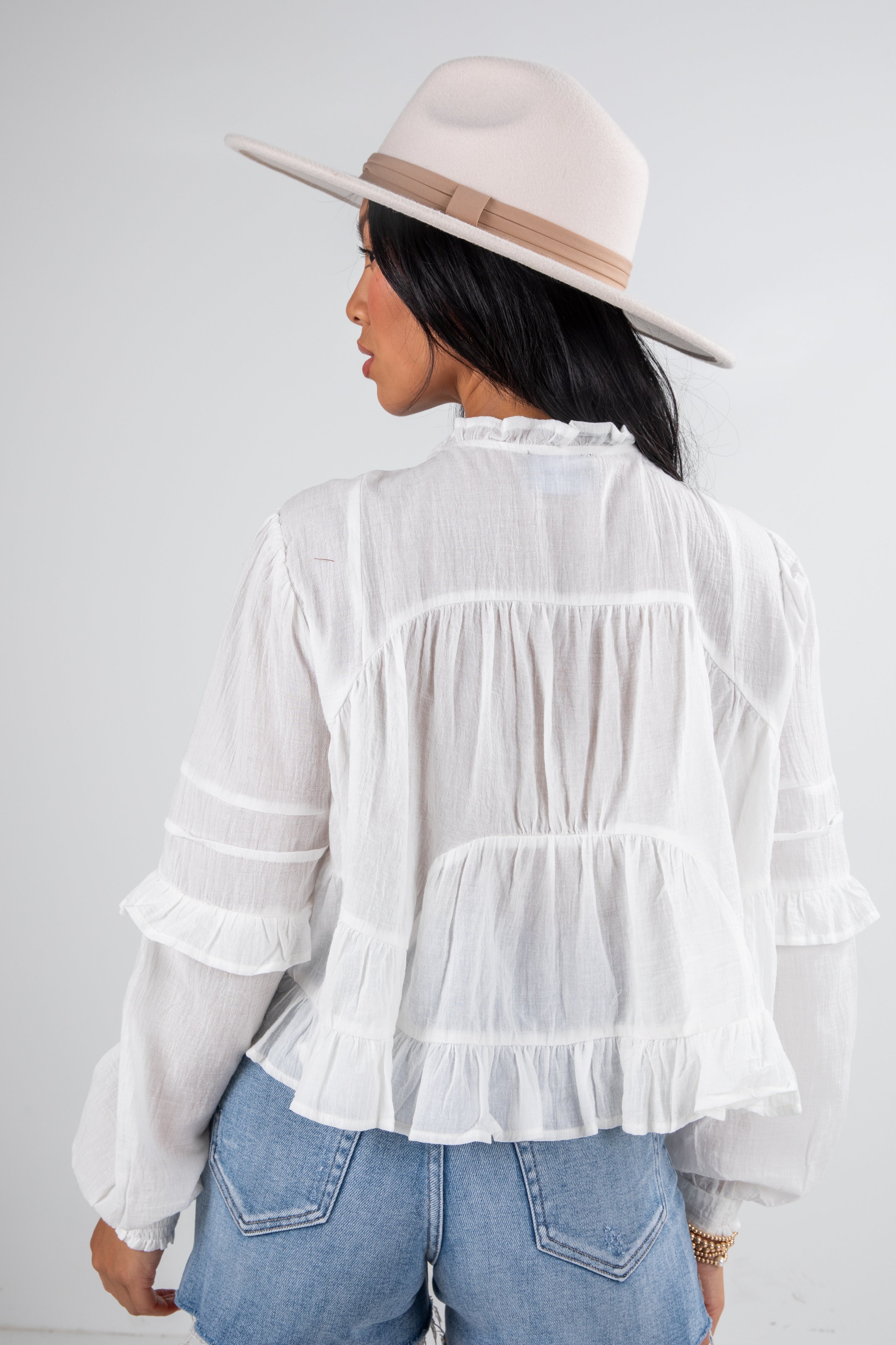 Person wearing a white ruffled blouse and blue jeans with a beige hat on a plain background