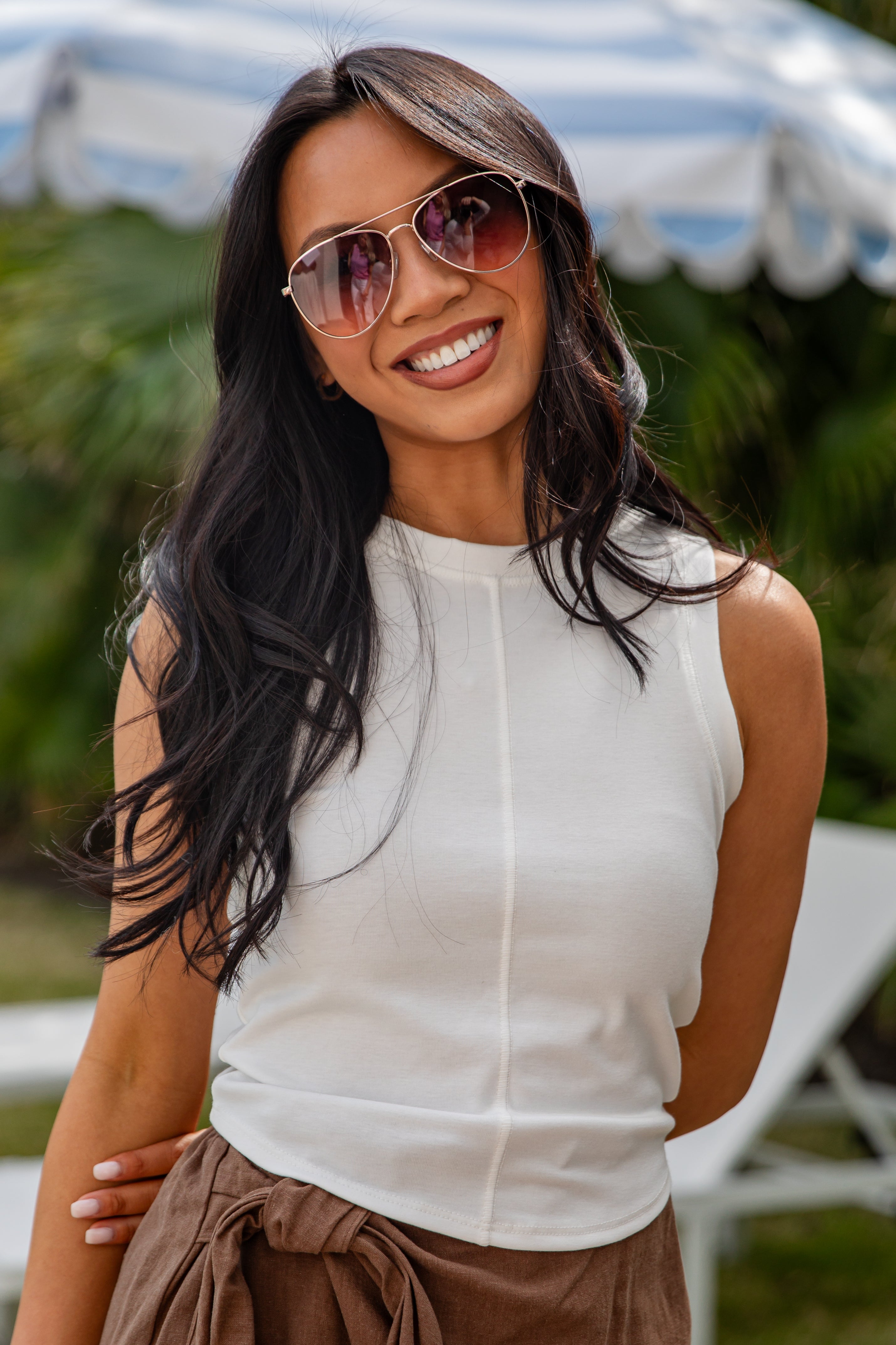 Woman wearing sunglasses and a white sleeveless top with a blurred outdoor background