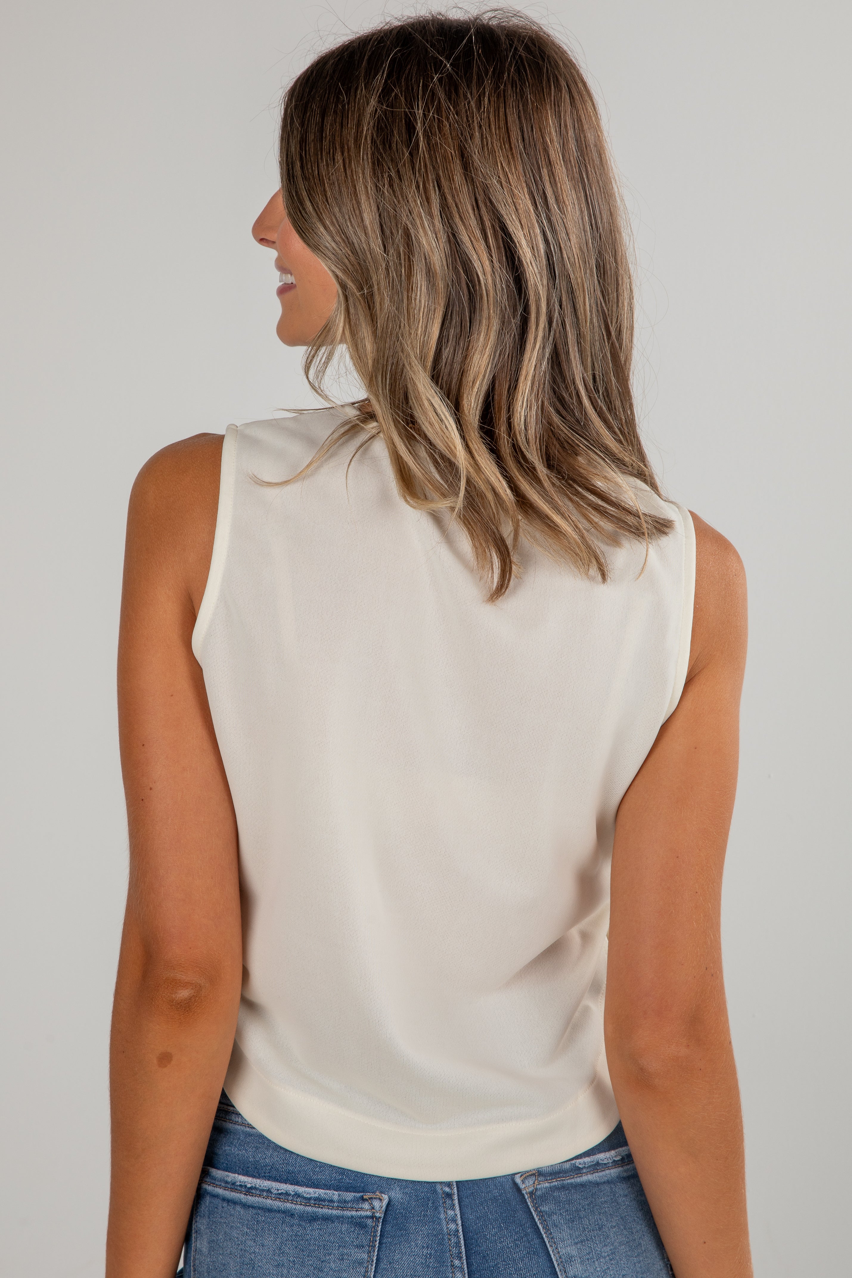 Woman wearing a beige sleeveless top and blue jeans against a plain background