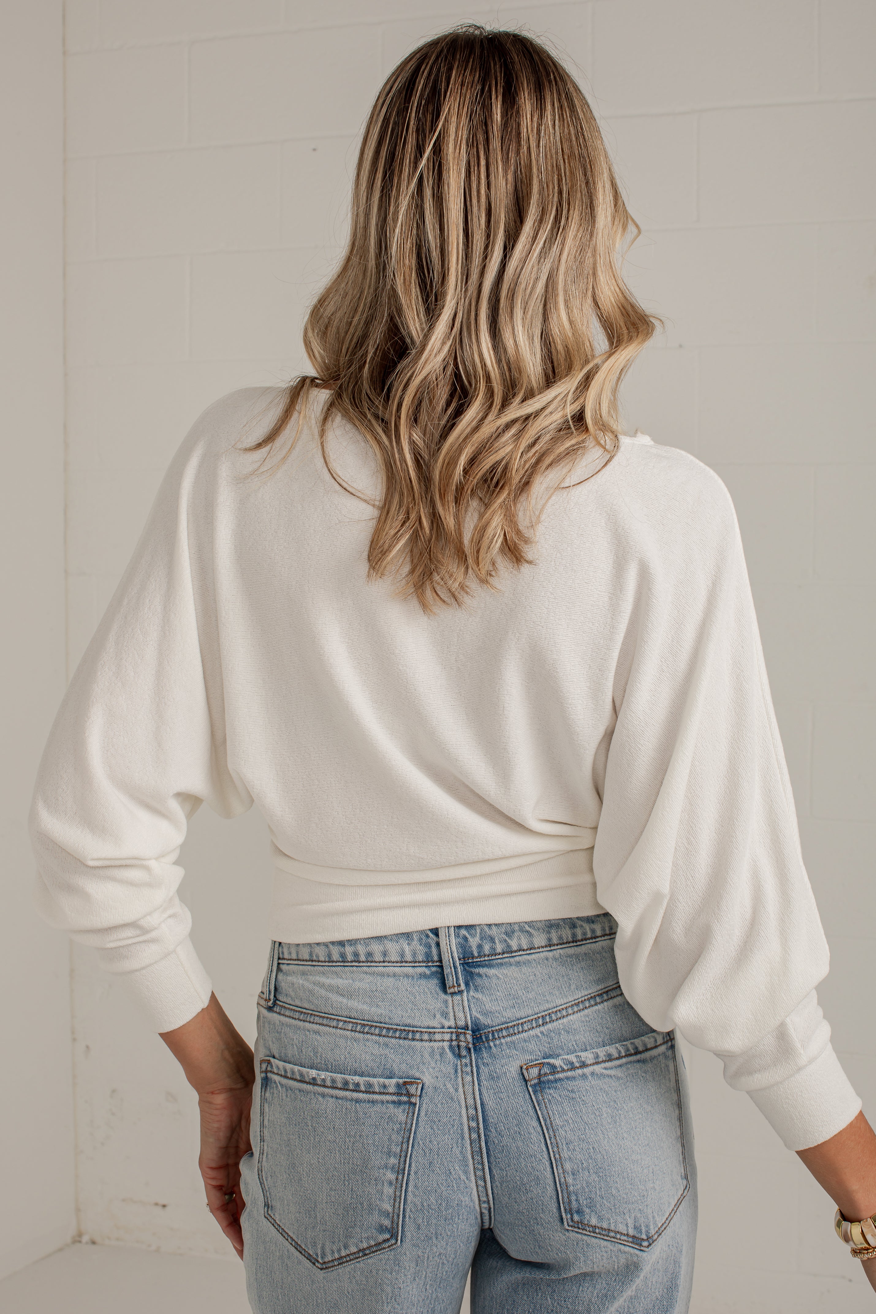 A woman viewed from behind wearing a cream-colored long-sleeve knit top and light-wash blue jeans, standing against a white tiled wall.