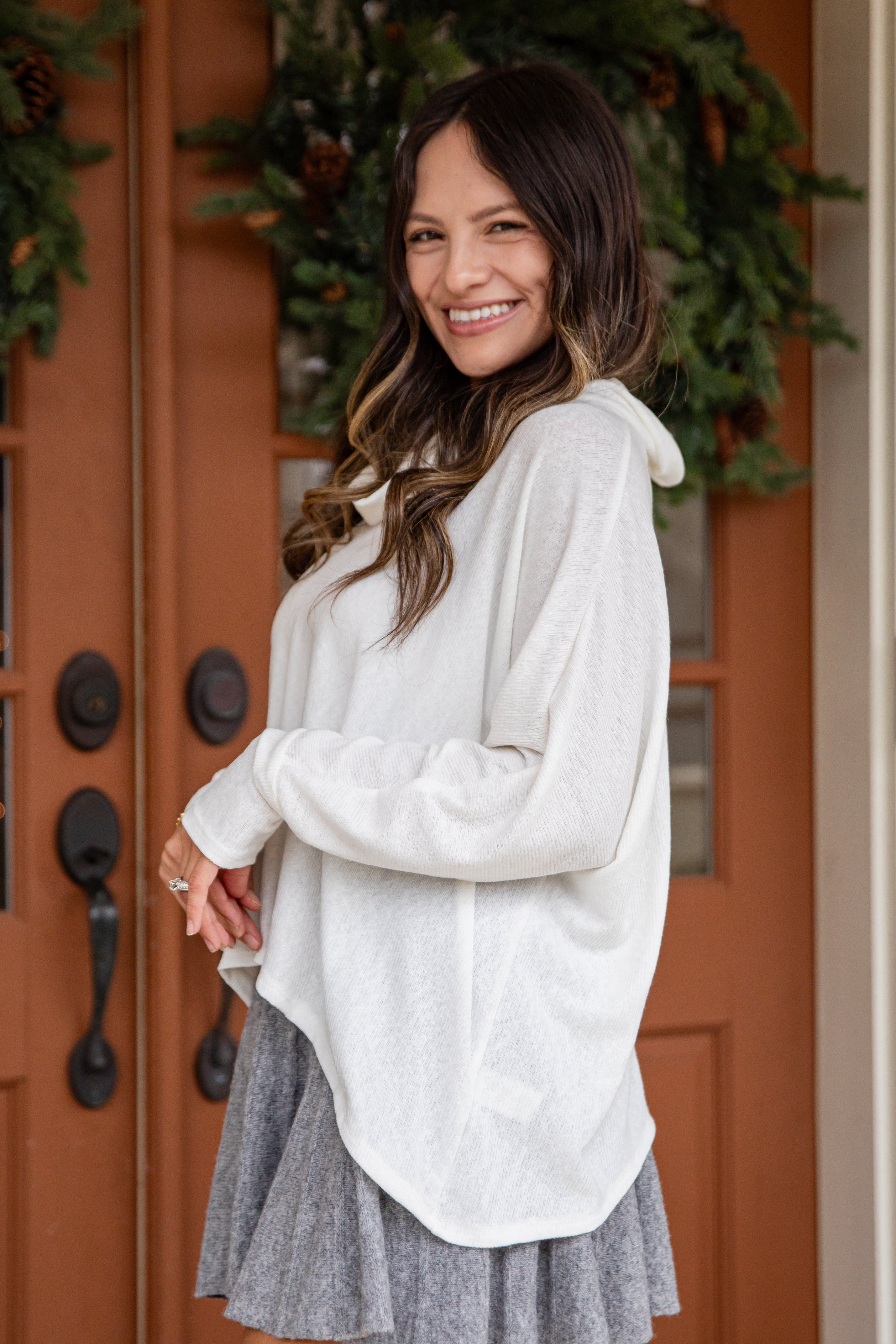 Woman wearing a light gray long-sleeve top and gray skirt standing in front of a wooden door with greenery.