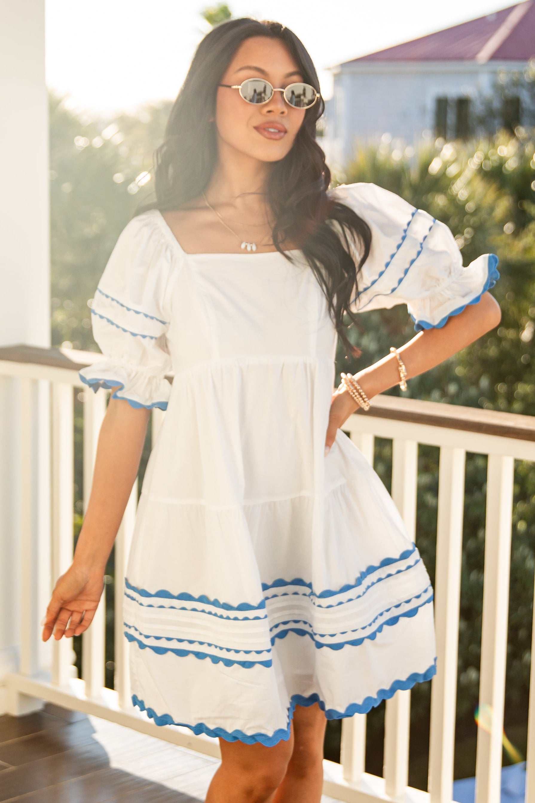 Woman in a white dress with blue accents standing on a balcony.