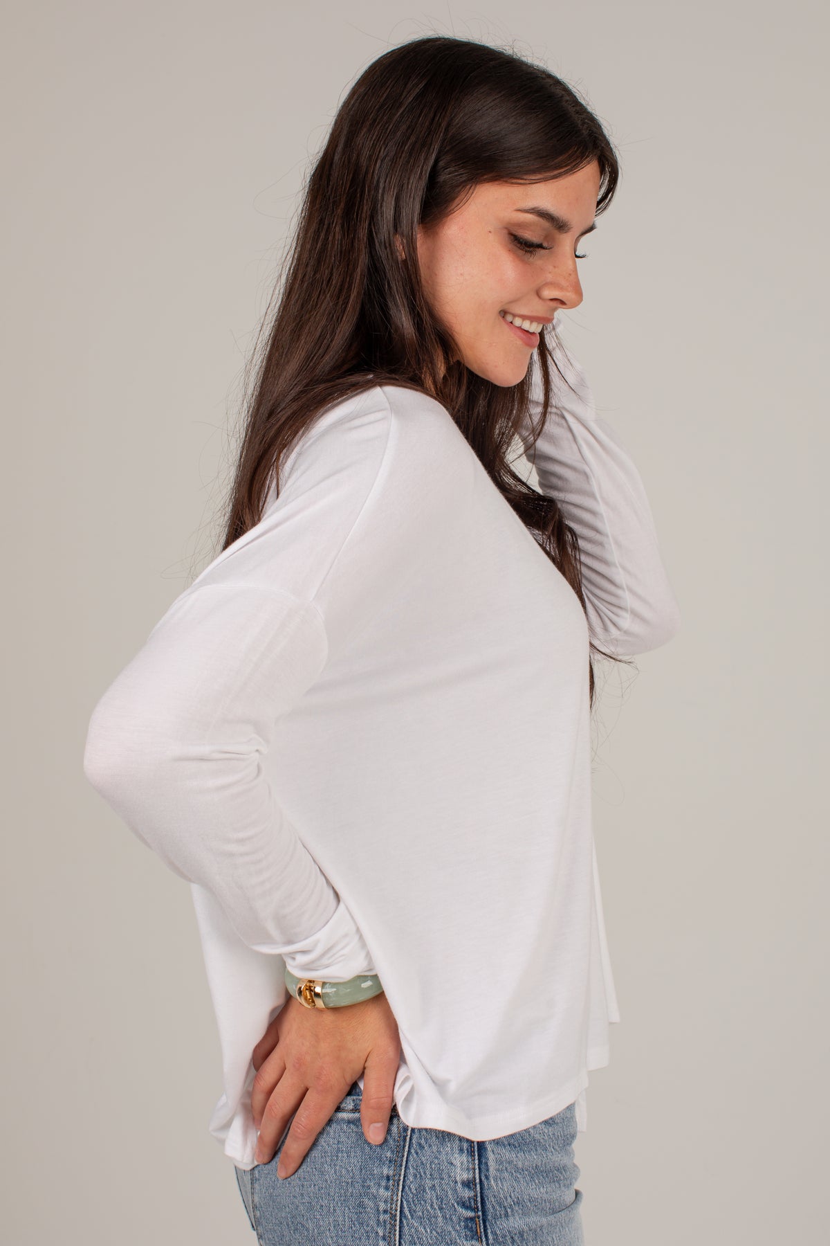 A woman wearing a white long-sleeve knit top and light blue jeans, shown from a side profile against a plain light gray studio background.