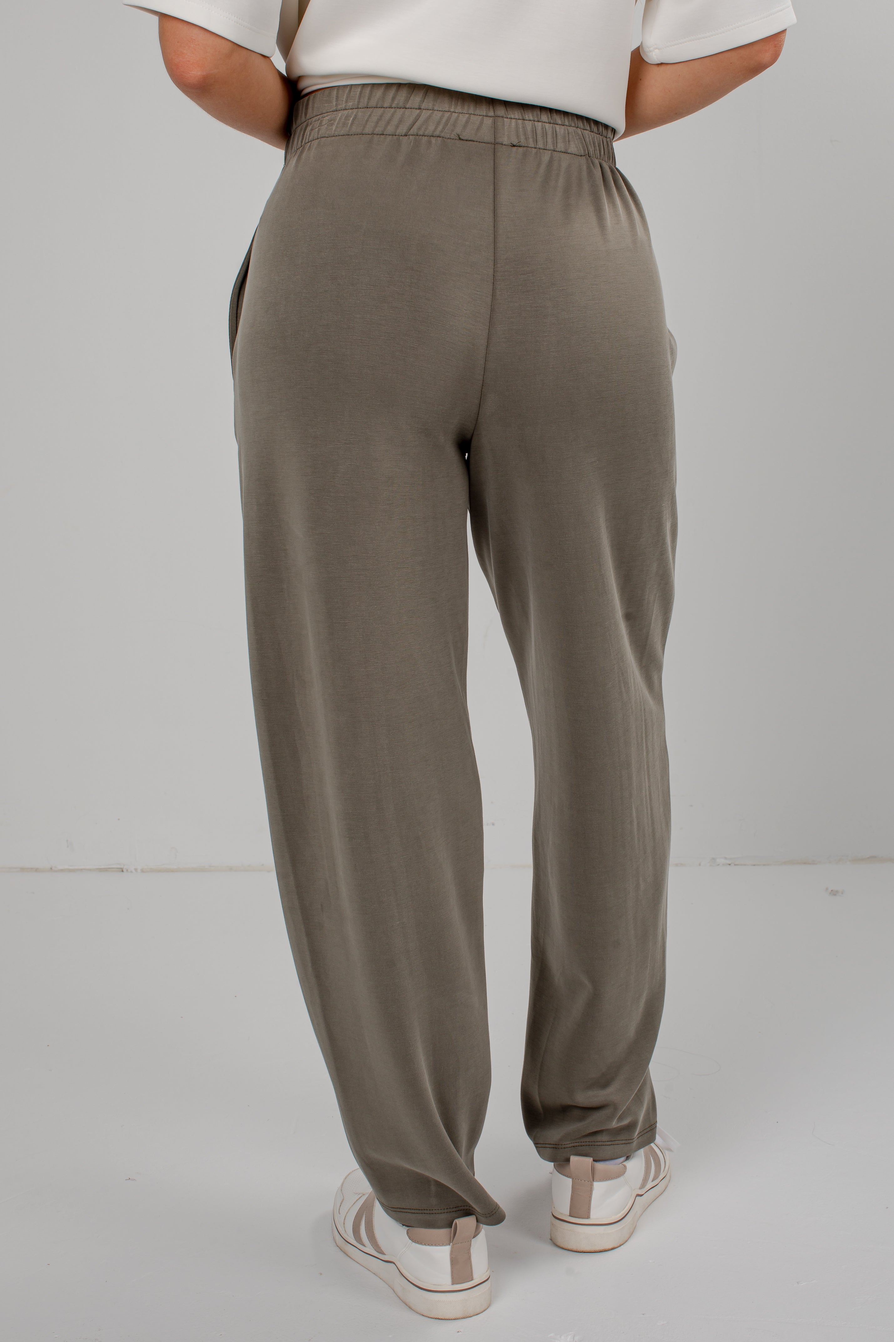 A person shown from the back wearing loose, wide-leg jogger-style pants in a muted taupe/olive-brown tone, paired with a white top and white sneakers, against a plain light gray studio background.