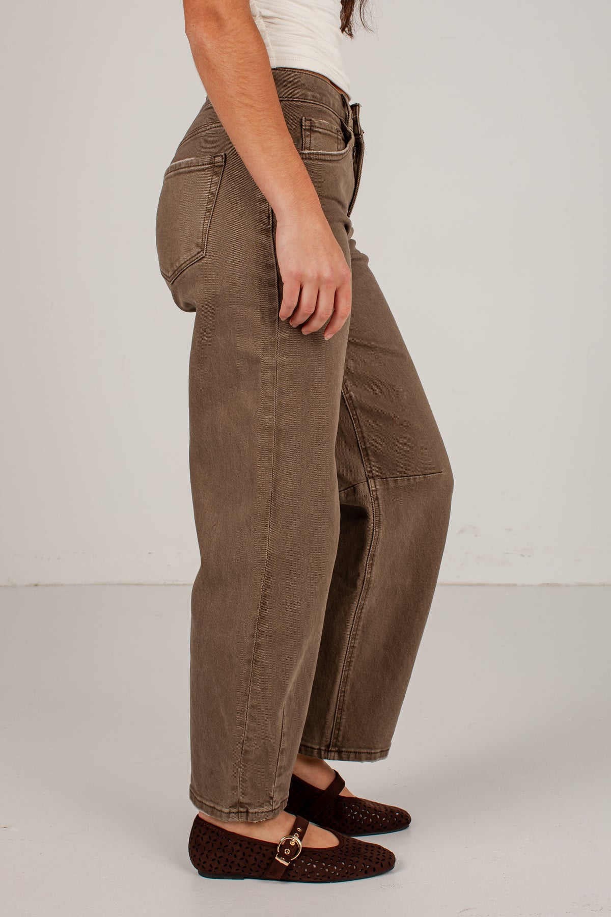 A side-profile view of a person wearing brown wide-leg cropped jeans, a white top, and dark brown woven flats against a plain light gray studio background.