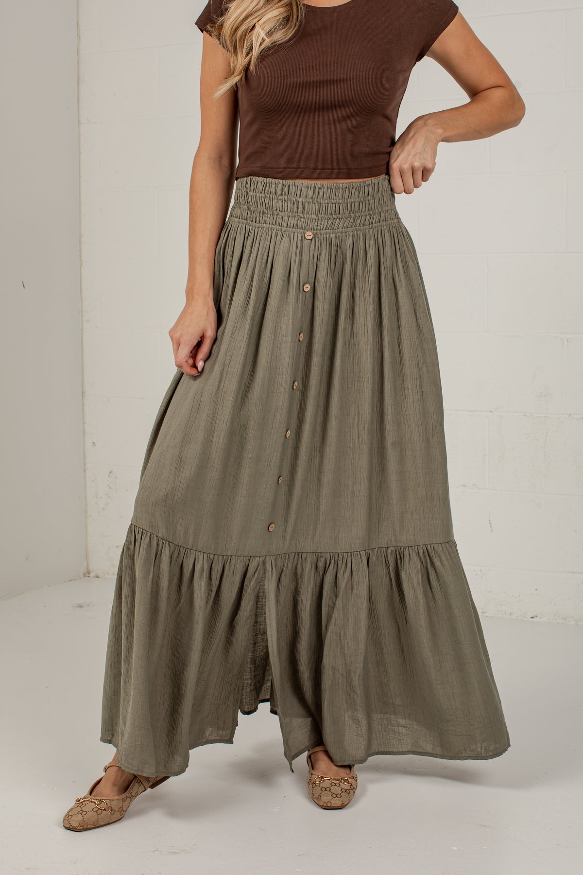 A woman wearing a long, tiered olive green skirt with a button-down front and a brown ribbed top, standing against a white studio wall.