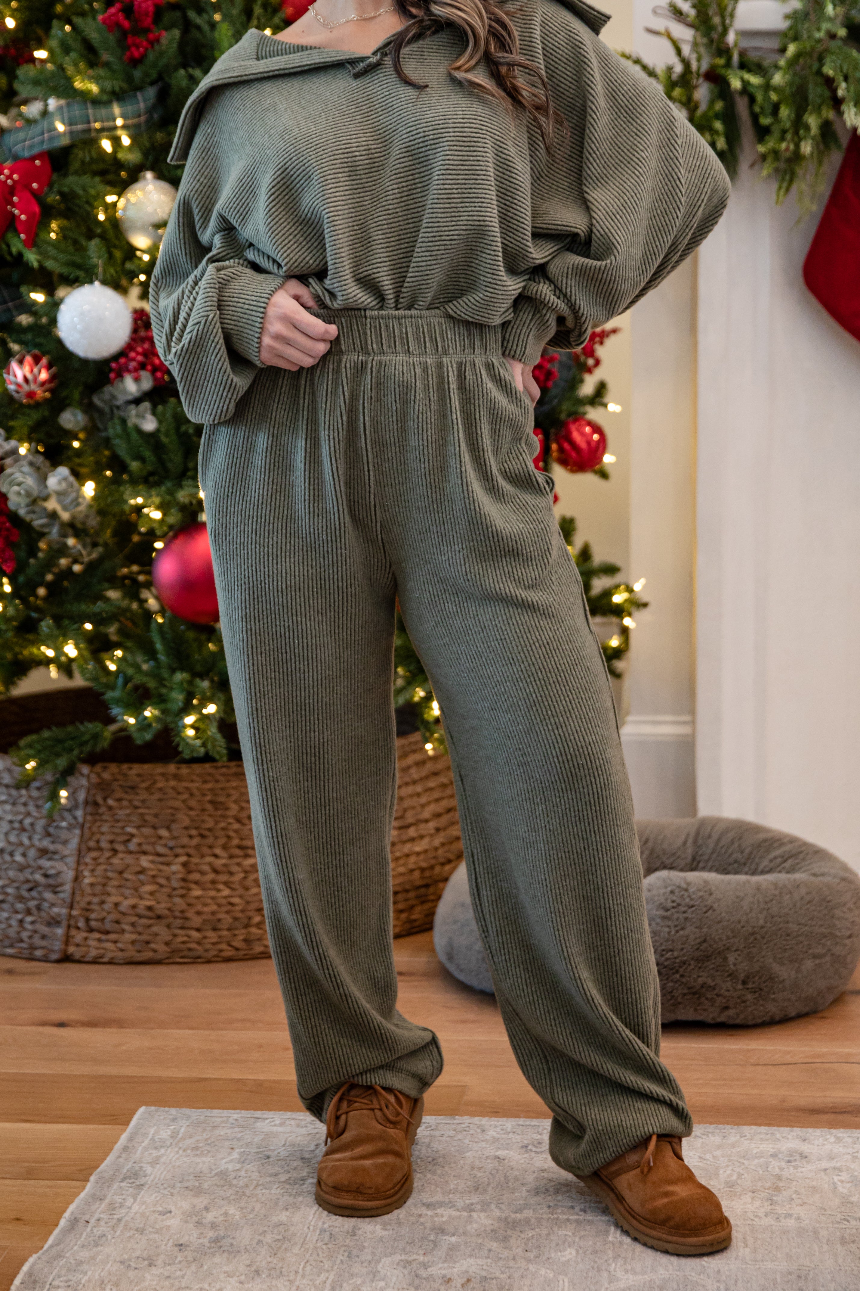 Person wearing a green ribbed sweater and pants set with brown shoes in front of a decorated Christmas tree.