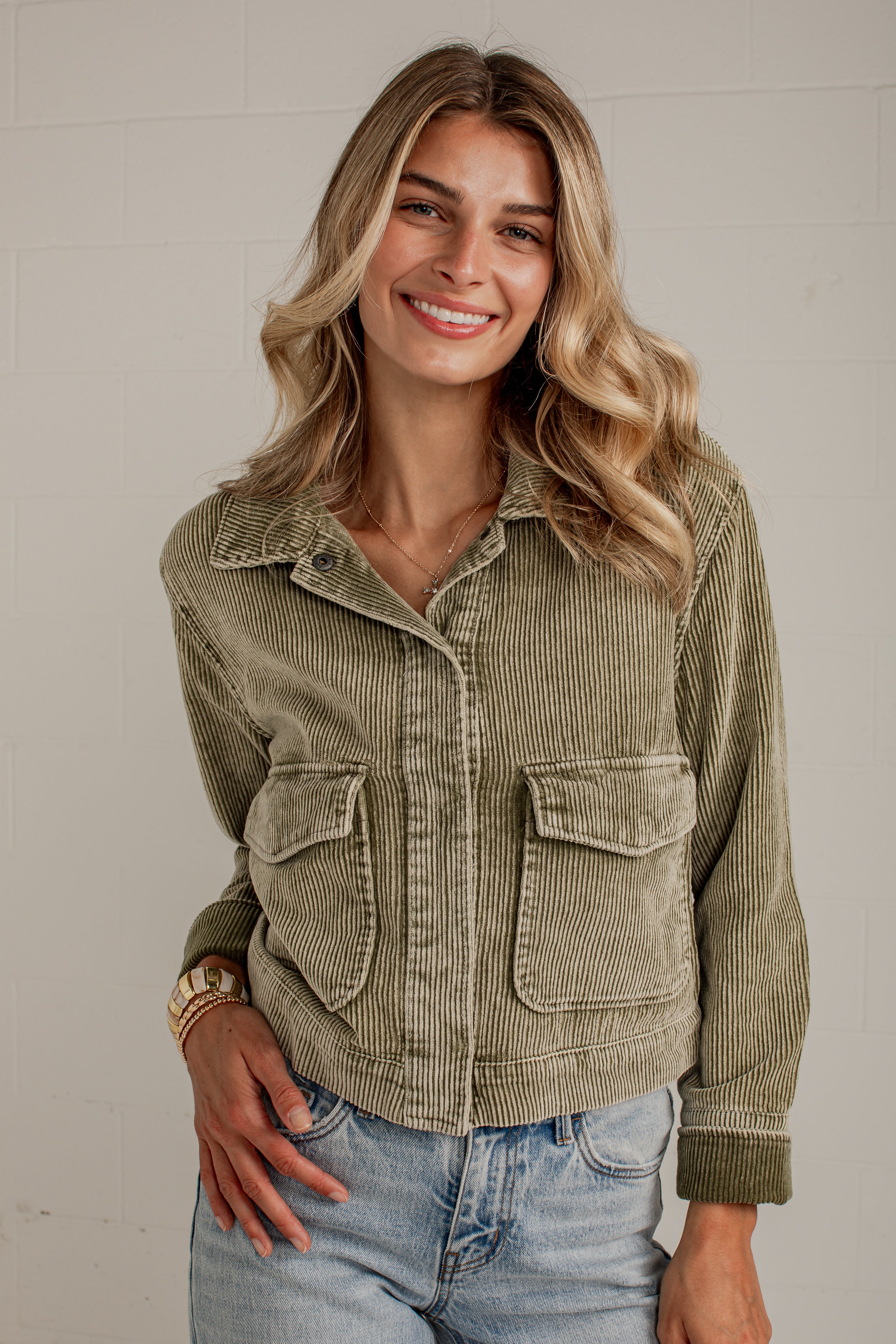 A woman wearing a cropped corduroy jacket in a muted sage green color, paired with light-wash blue jeans, standing against a light-colored brick wall.