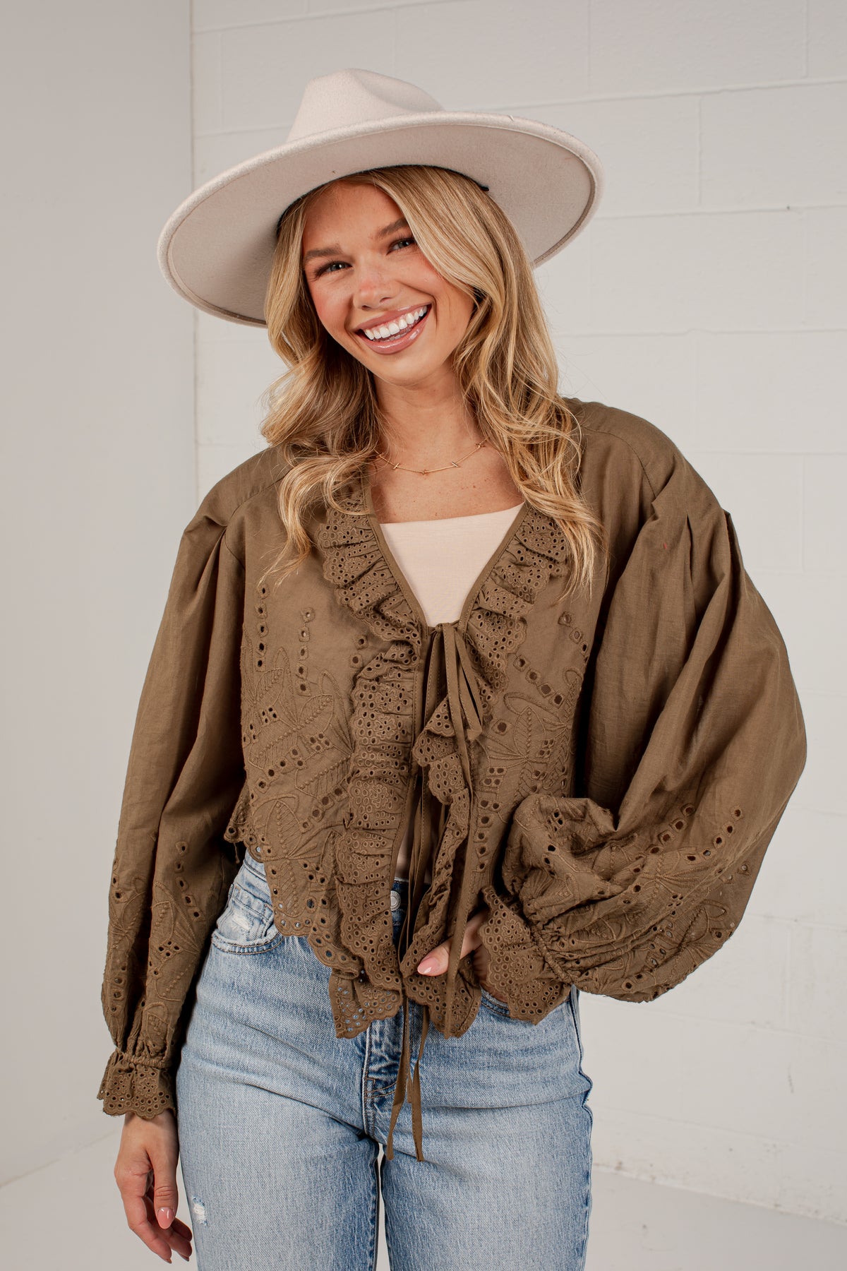A woman wearing a brown eyelet lace blouse with ruffles and tie-front detailing, styled with a beige wide-brim hat, a cream top, and light-wash blue jeans against a white brick wall.