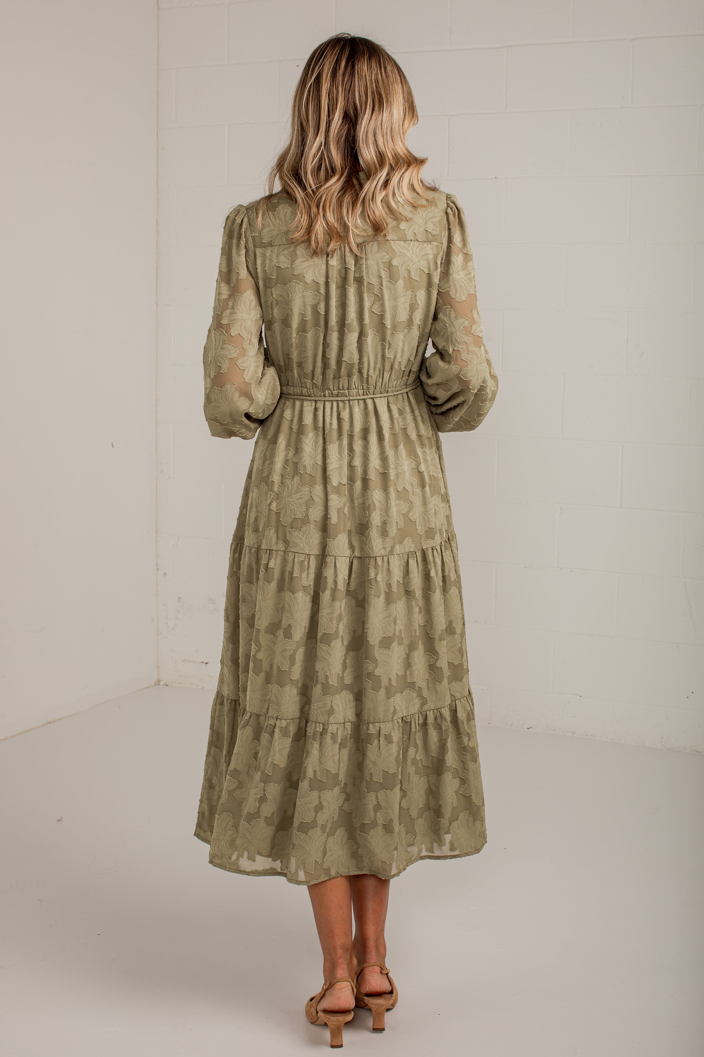 A woman viewed from behind wearing a long-sleeve, tiered midi dress in a muted olive green and beige patterned fabric, paired with tan heeled sandals, set against a plain white studio background.