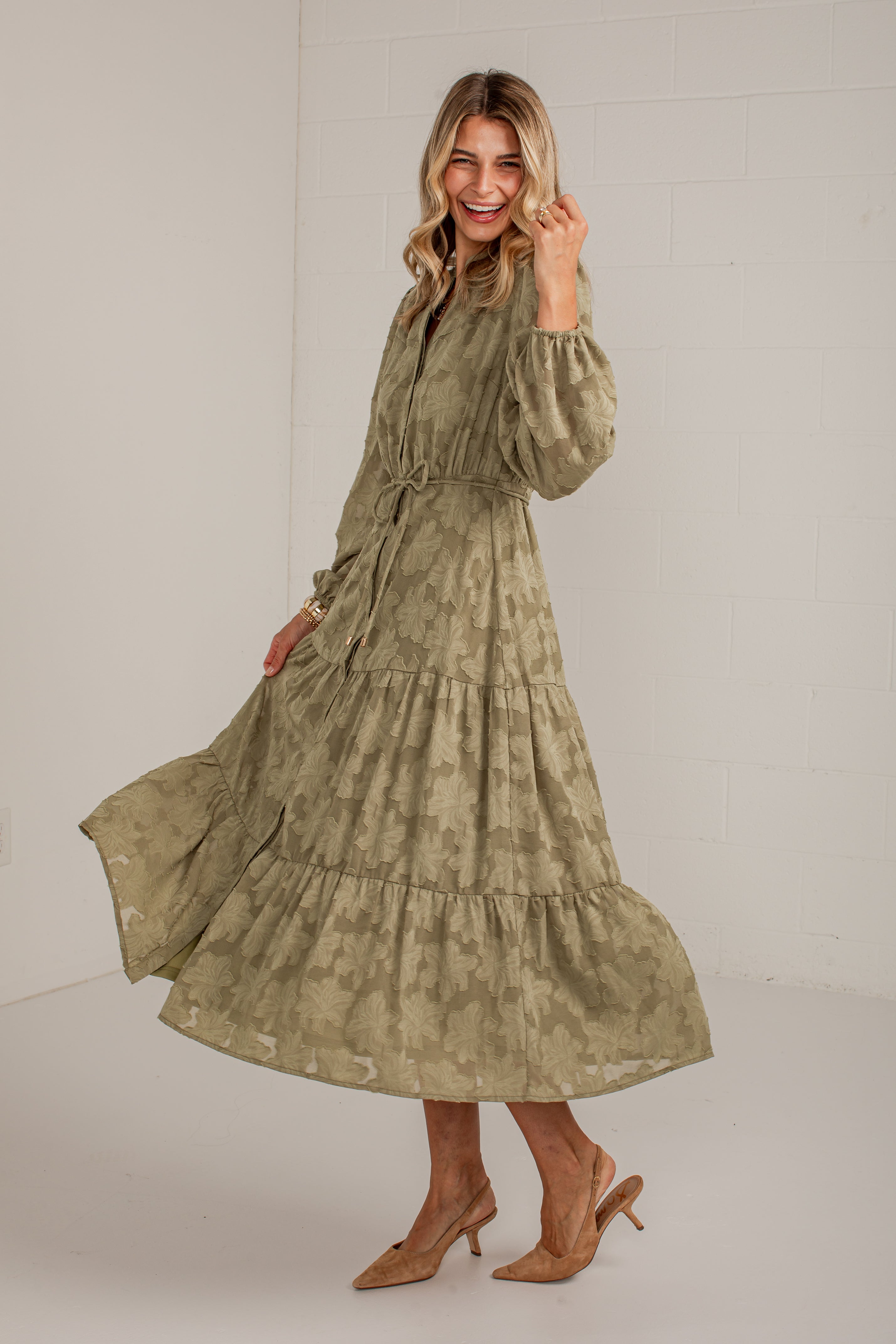 A woman modeling a long-sleeved, tiered midi dress with a muted olive green and beige camouflage pattern, paired with tan heeled sandals, set against a white studio background.
