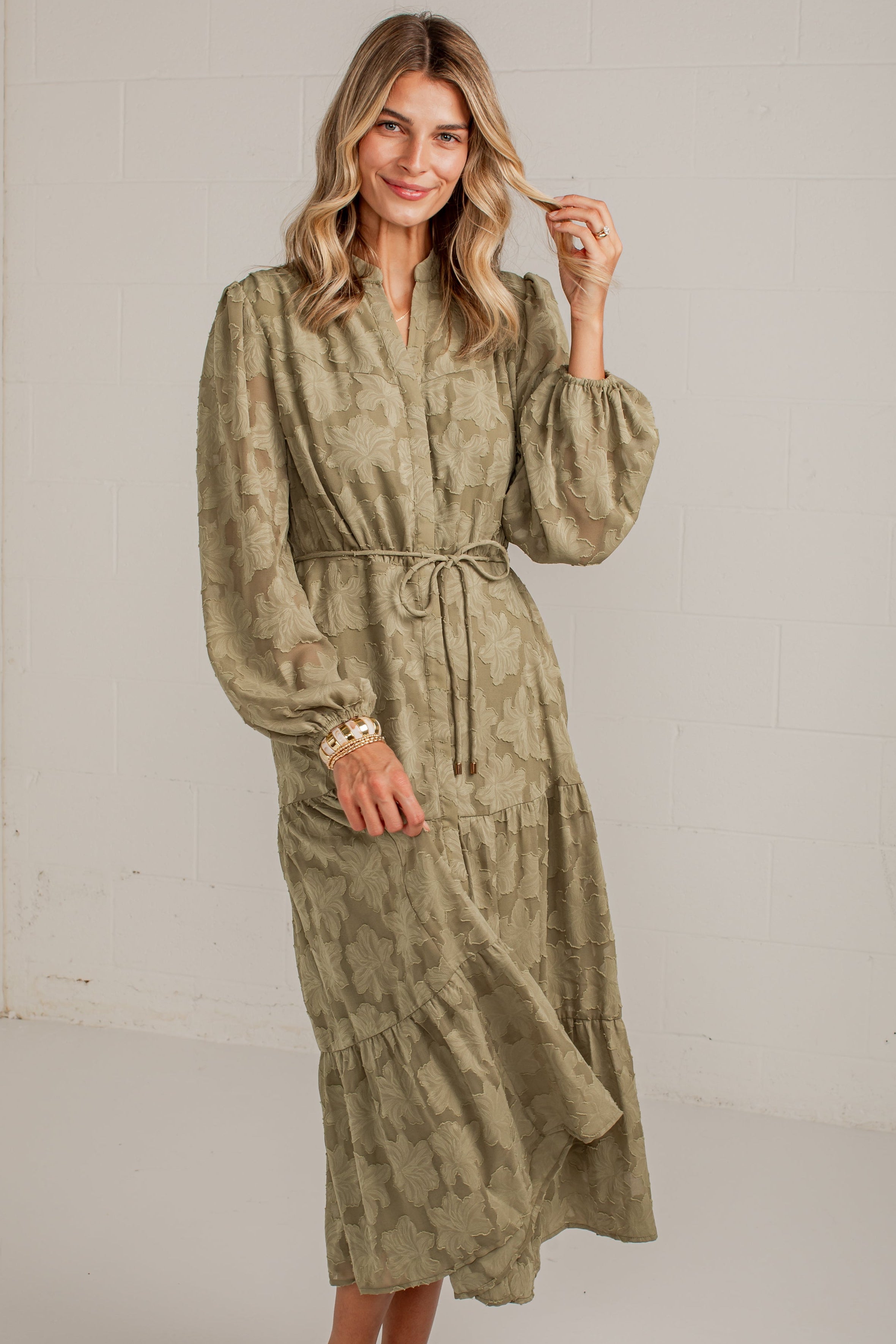 A woman modeling a long-sleeved olive green and beige patterned midi dress with a tie waist, standing in a bright white studio setting.
