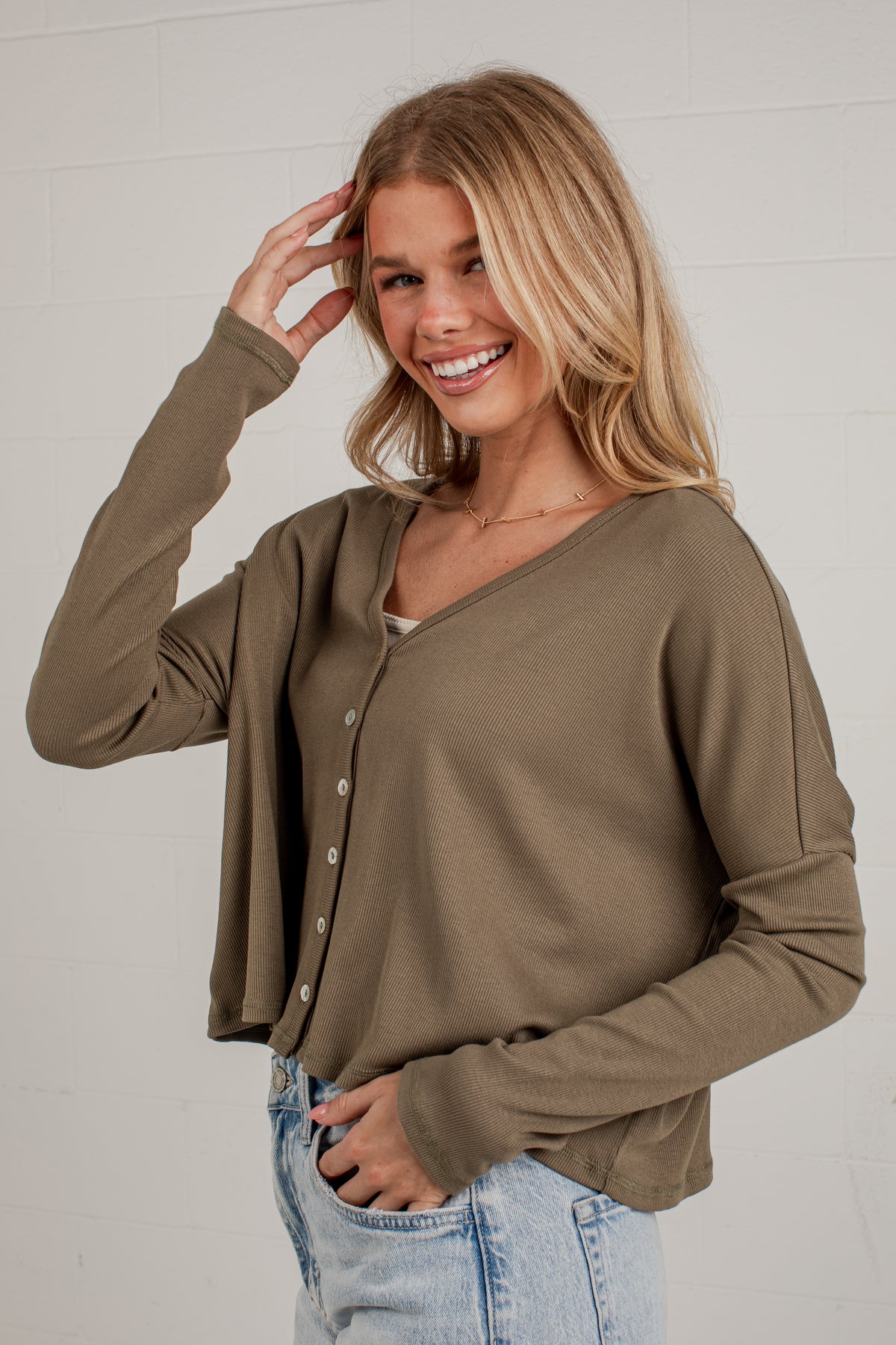 A woman wearing a long-sleeve V-neck button-front top in a muted olive-brown color, paired with light-wash blue jeans, standing against a light gray brick wall.