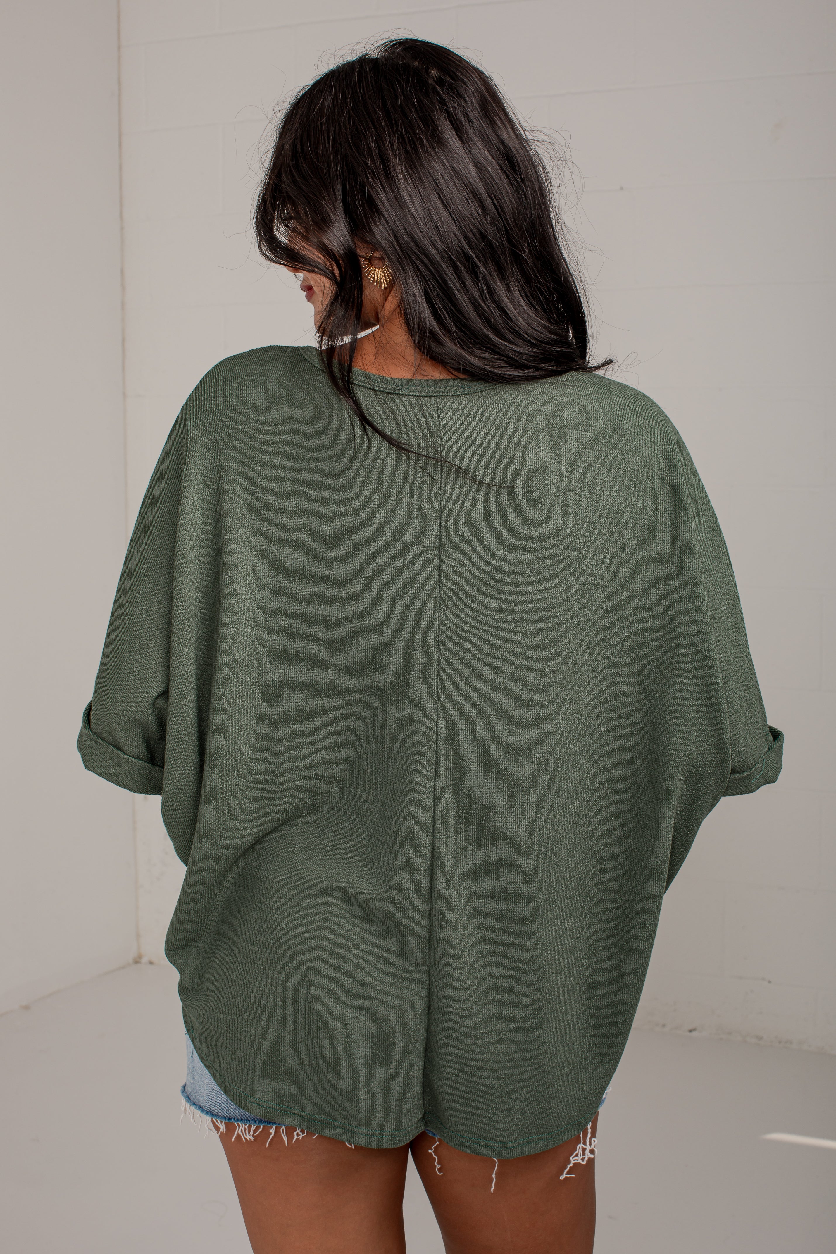 A woman viewed from behind wearing an oversized sage green knit top with rolled sleeves and a split back seam, paired with denim shorts.