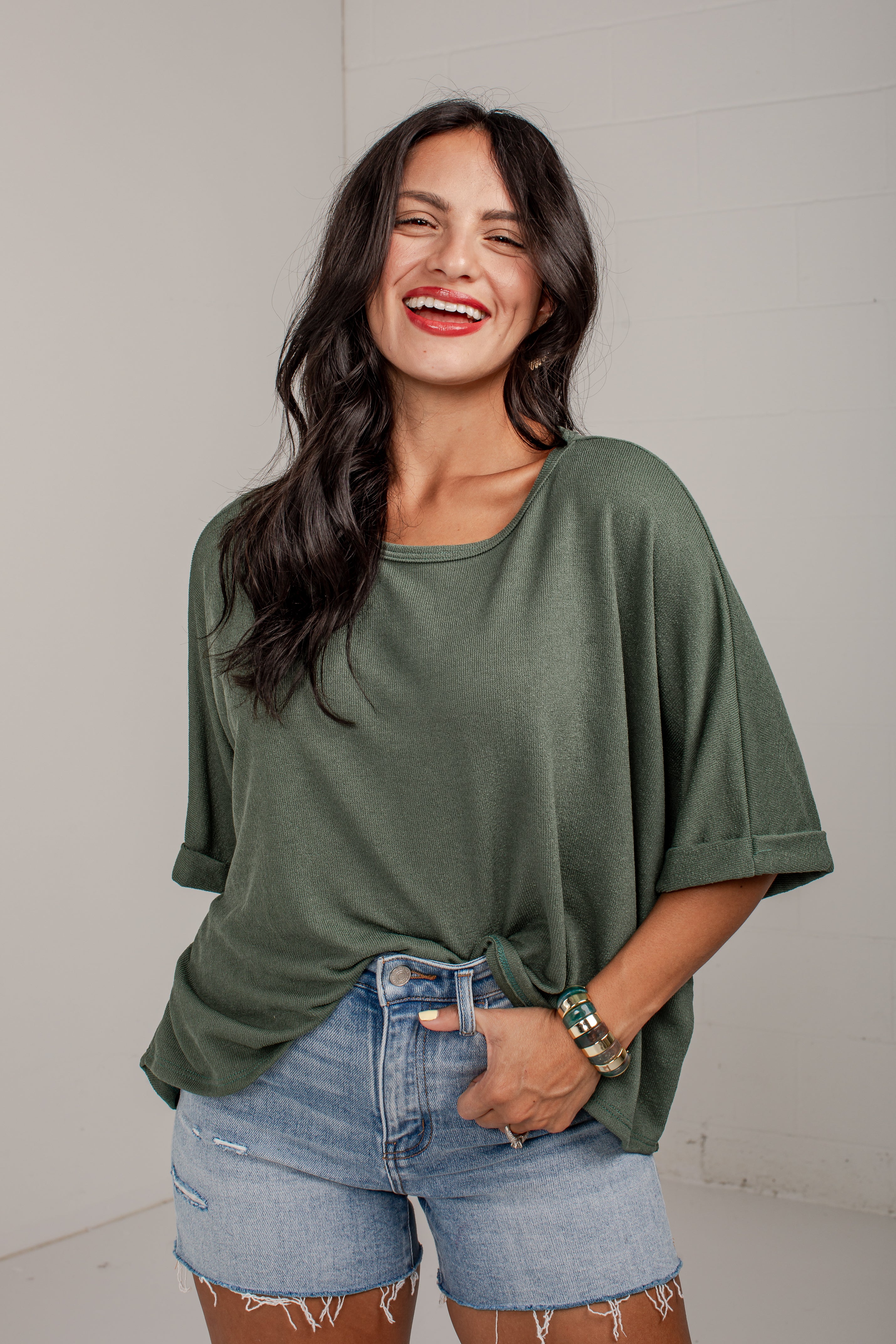 A woman smiling and wearing an olive green short-sleeve tunic top with light blue denim shorts, standing against a light gray wall.