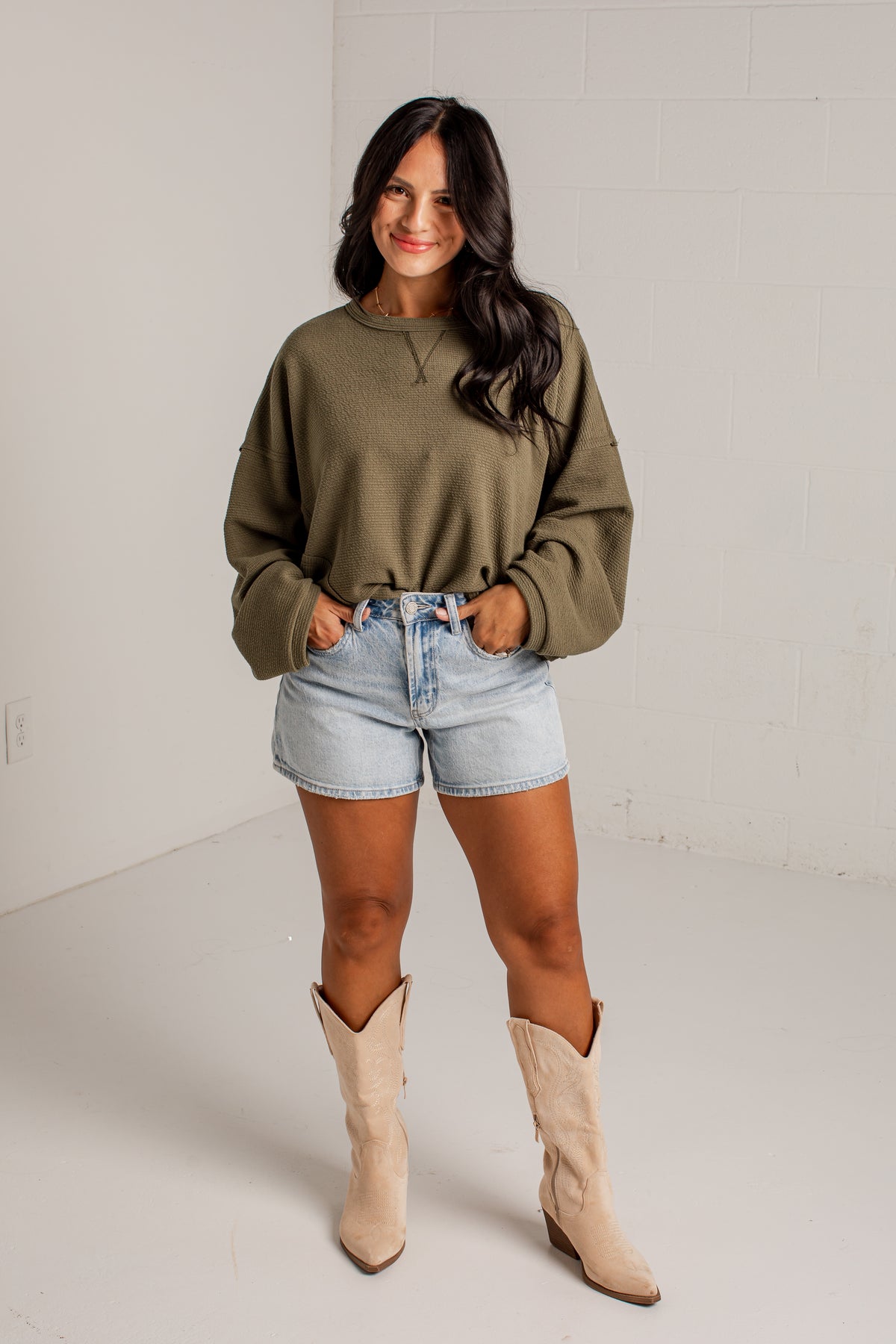 A woman modeling a ribbed crewneck sweatshirt in olive green, paired with light-wash denim shorts and beige cowboy boots, standing in a bright white studio setting.