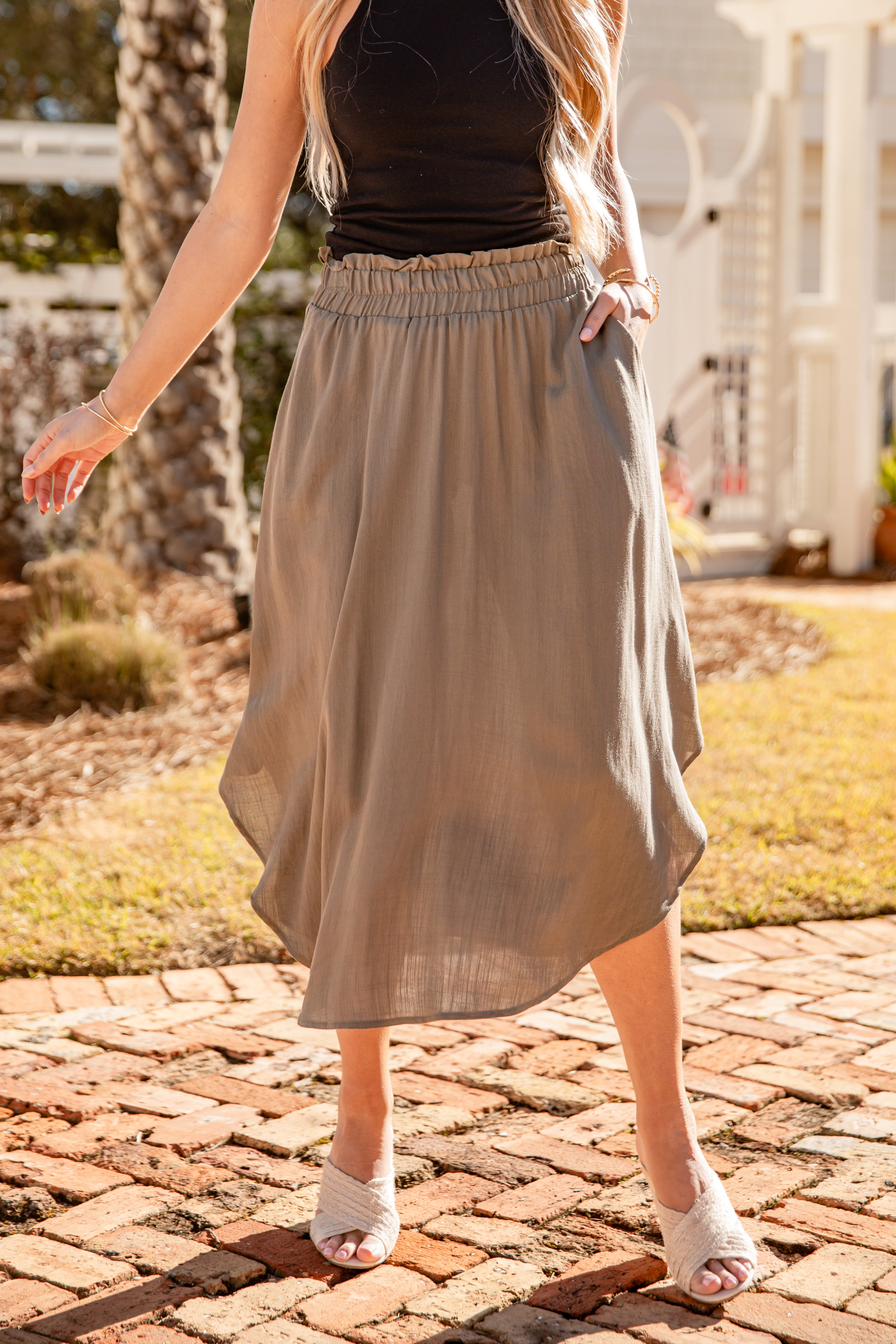 Woman wearing a beige skirt and black top on a brick path with greenery in the background
