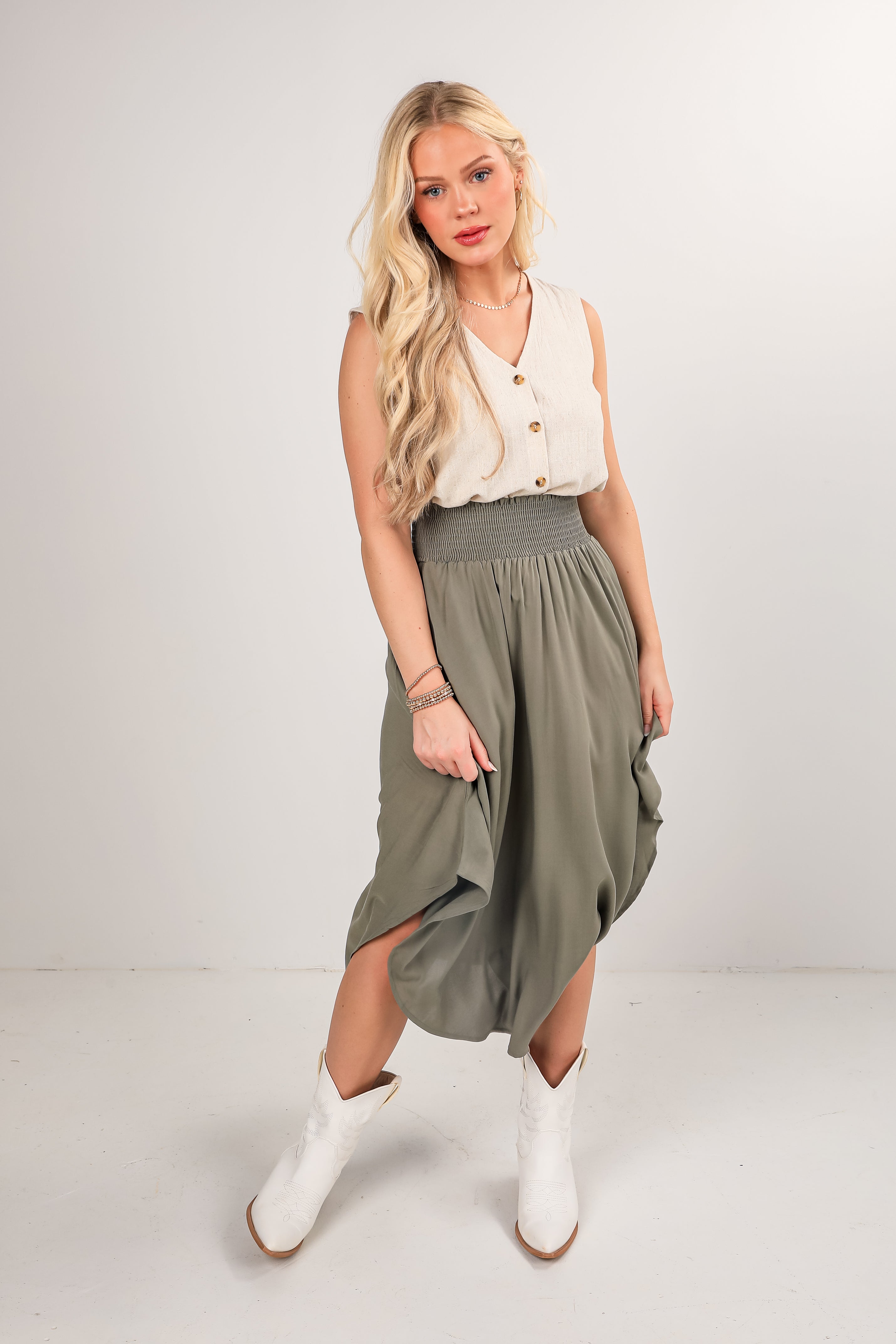 Woman wearing a white sleeveless top and green skirt with white boots on a plain background