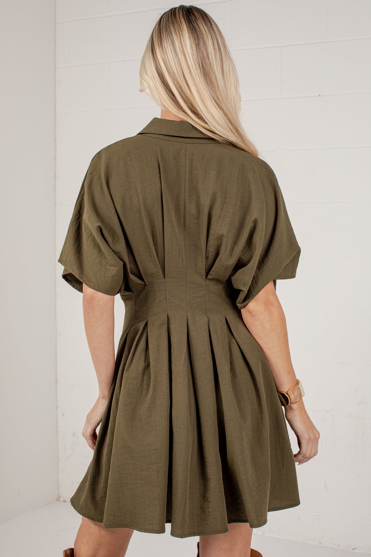 A woman viewed from behind wearing an olive green short-sleeve shirt dress with a pleated back and flared skirt, standing against a white paneled wall.