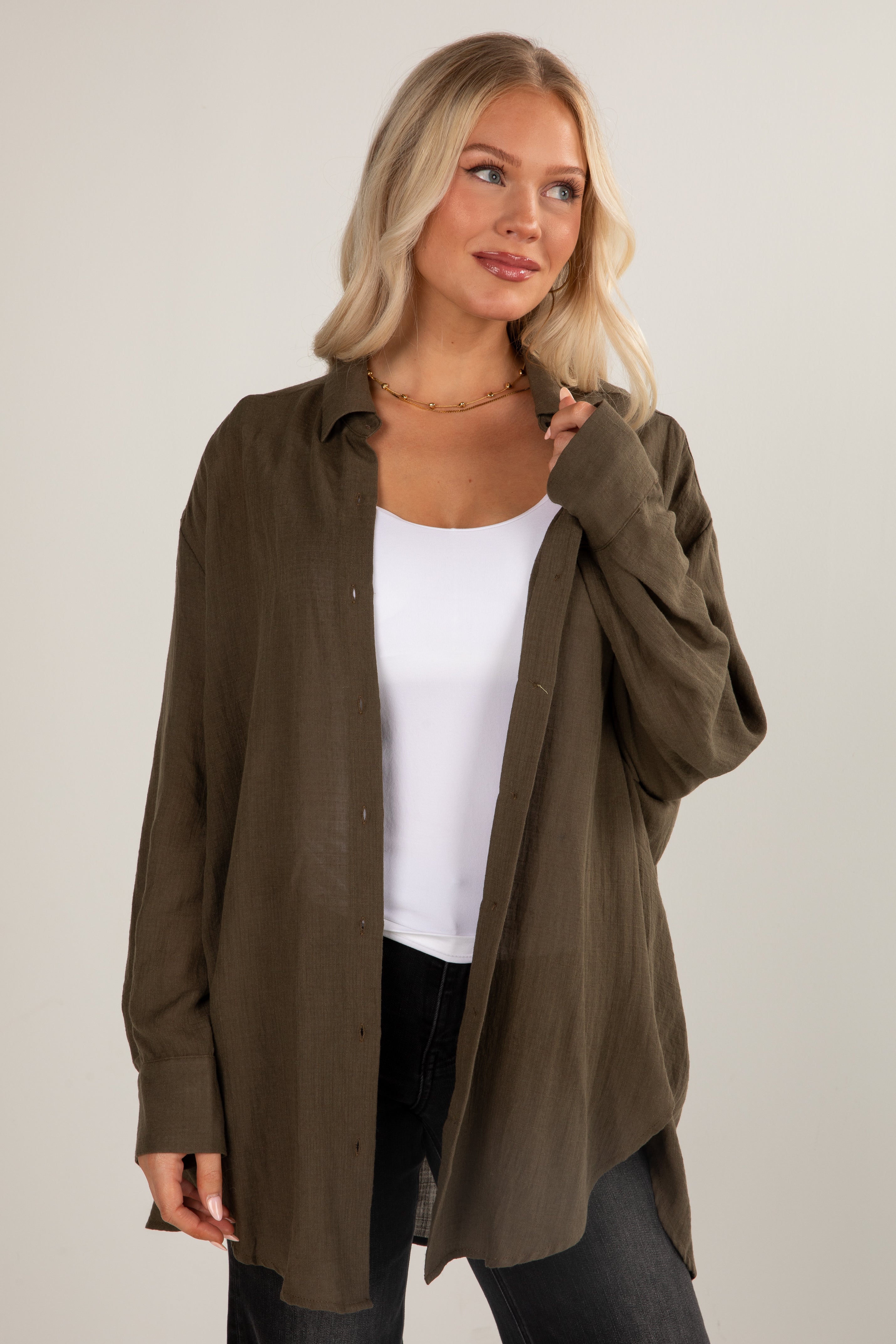 Woman wearing an olive green shirt over a white top and black pants on a plain background