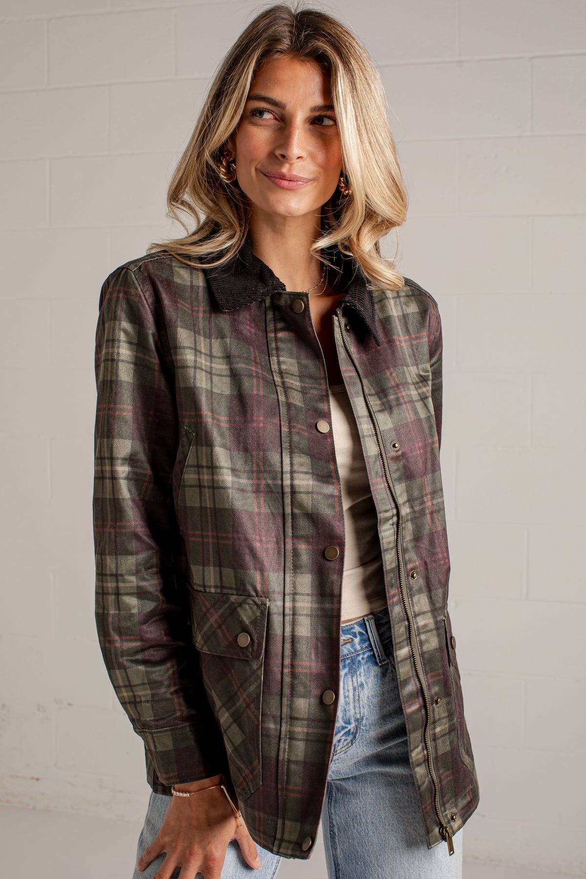A woman wearing a dark olive green and brown plaid button-up jacket over a beige top and blue jeans, standing against a white brick wall.
