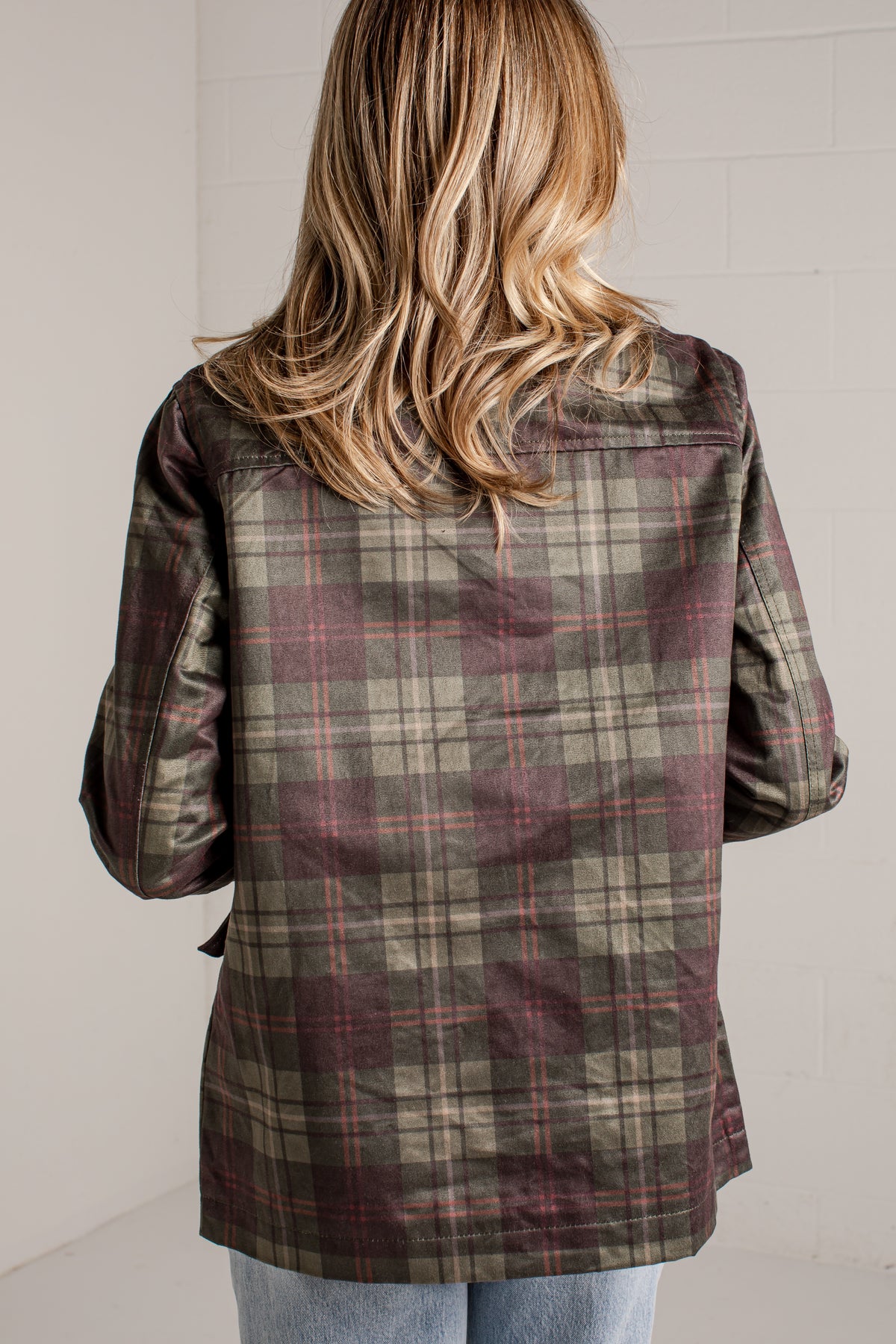 A woman viewed from behind wearing a long-sleeve plaid button-up shirt in olive green, brown, and burgundy tones, paired with light blue jeans against a white brick wall.