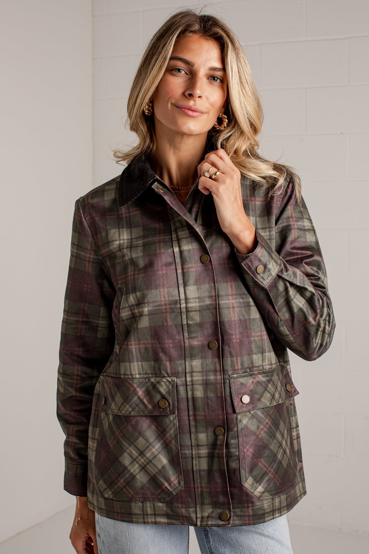 A woman wearing a brown and green plaid waxed jacket with dark brown corduroy collar and front buttons, paired with light blue jeans, standing against a white brick wall.