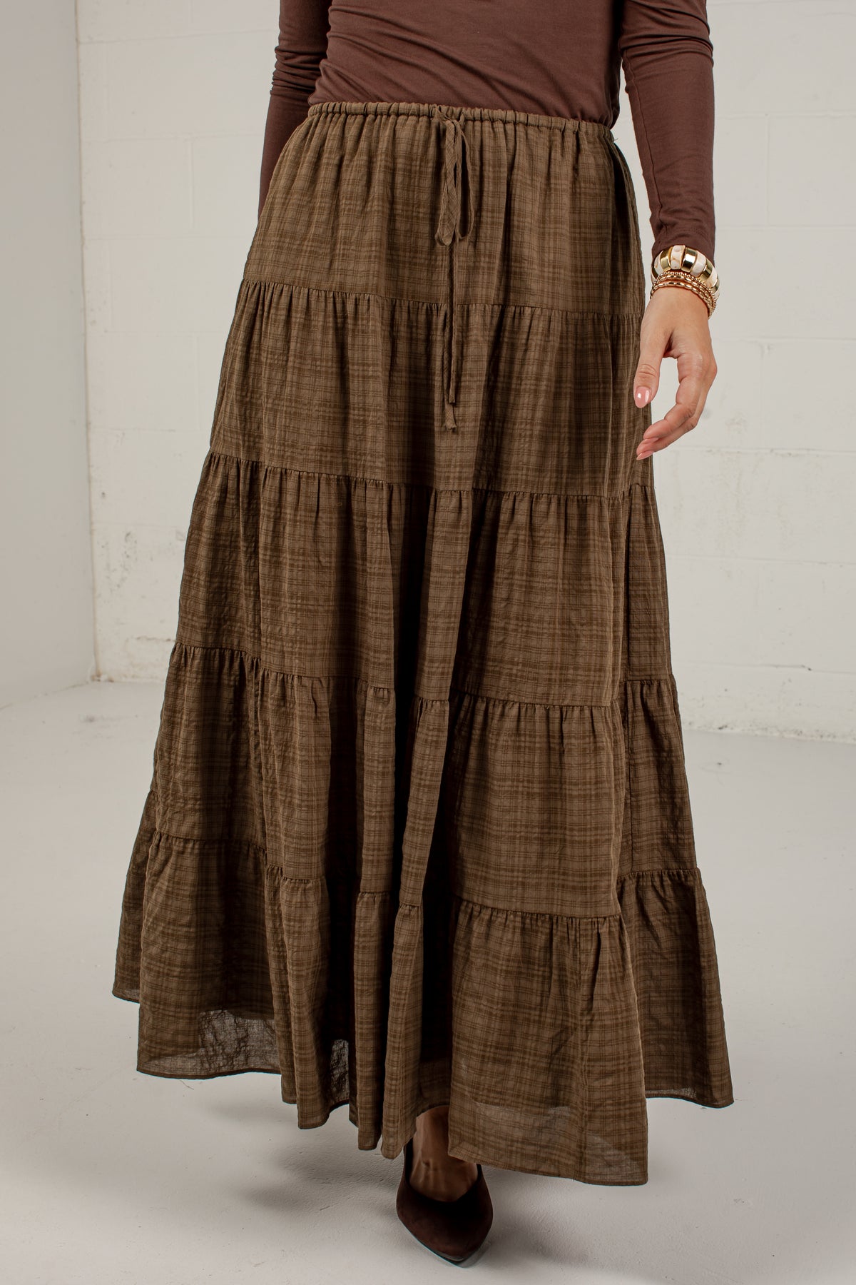 A brown tiered maxi skirt with a subtle plaid pattern, worn by a model with a brown long-sleeve top and dark brown pointed-toe heels.