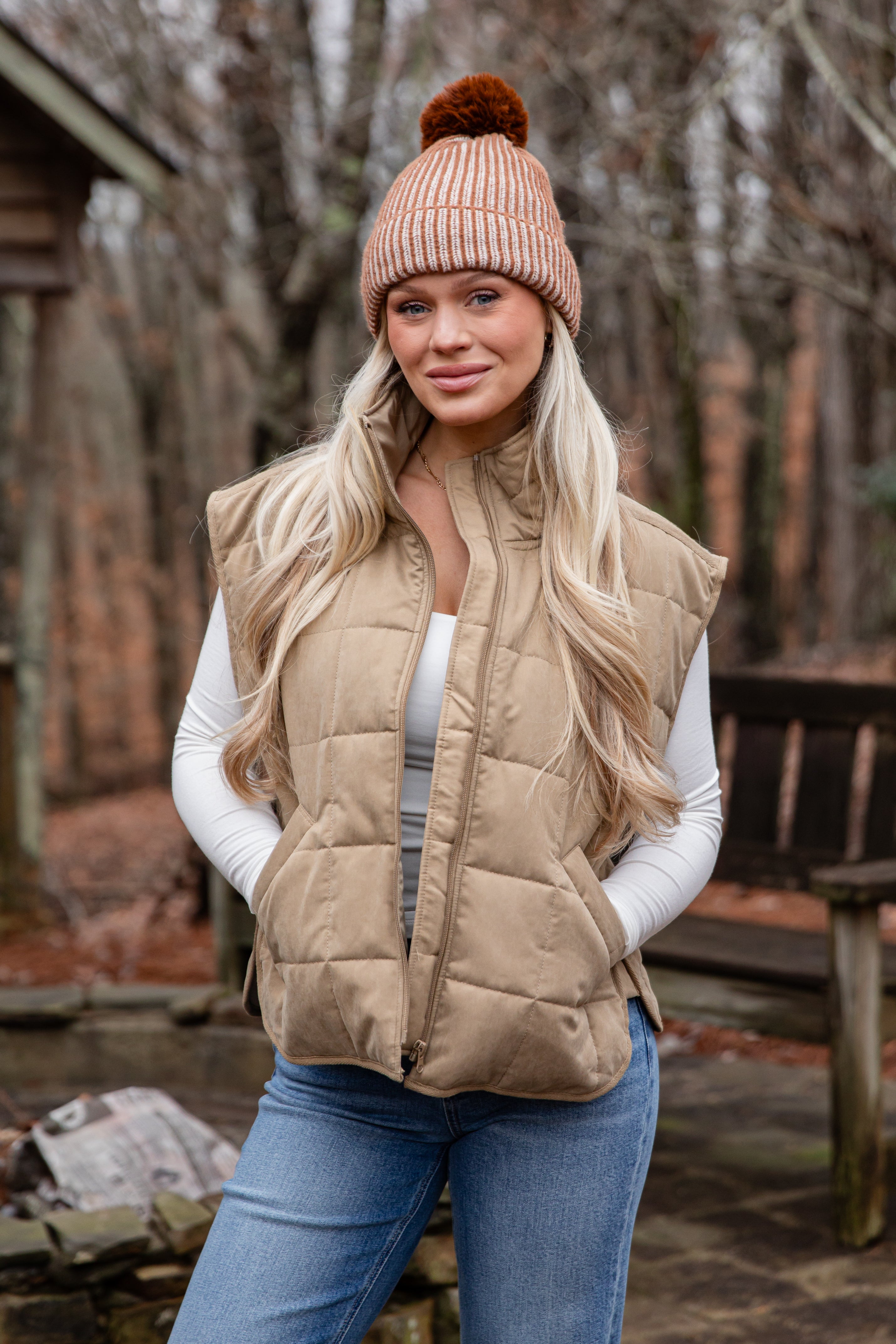 Woman wearing a beige puffer vest, white sweater, blue jeans, and a matching beanie outdoors.