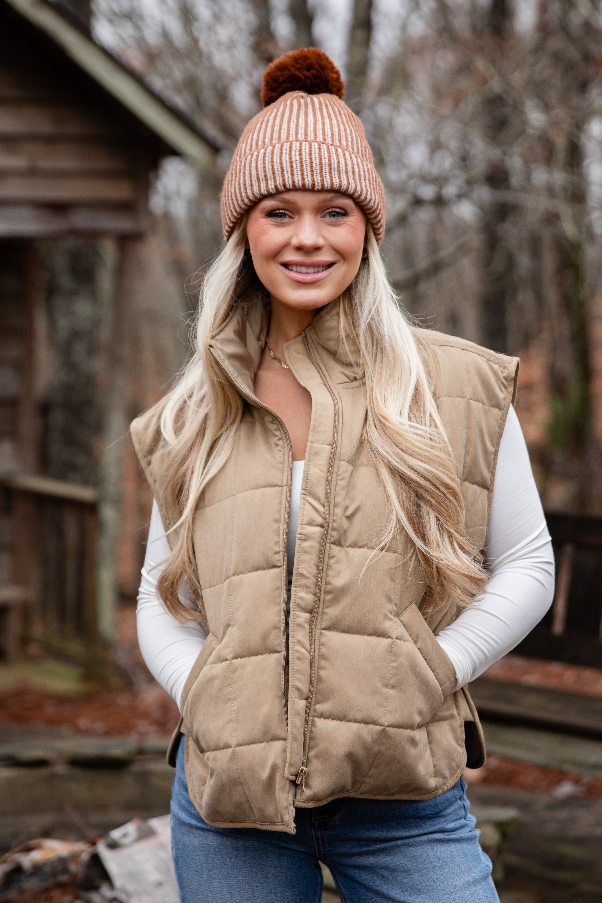Woman wearing a beige puffer vest and beanie outdoors near a wooden cabin.