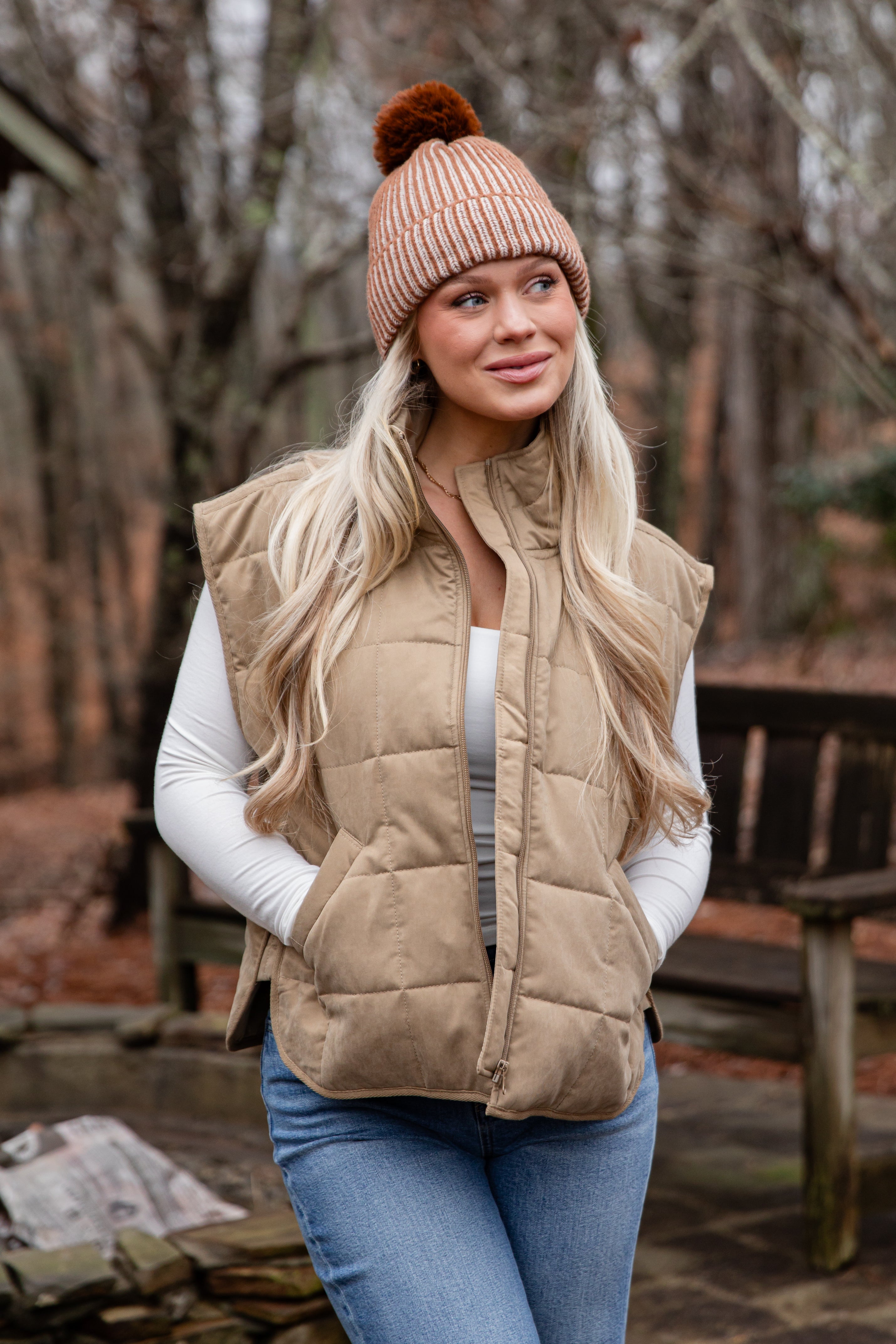 Woman wearing a beige puffer vest, white sweater, blue jeans, and a beige knit hat with a pom-pom in a forest setting.