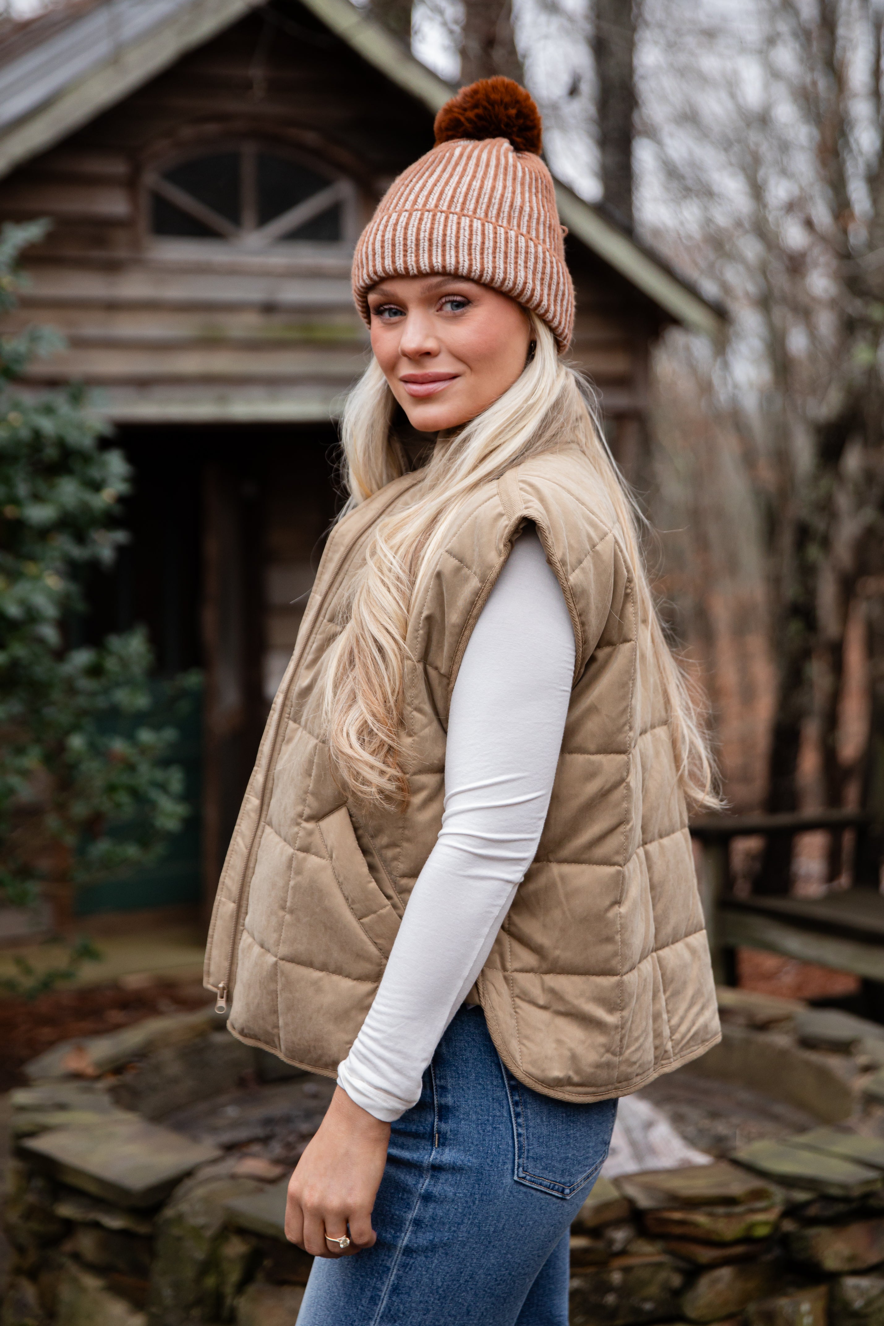 Woman wearing a beige puffer vest, white long-sleeve shirt, blue jeans, and a matching beanie in front of a wooden cabin.