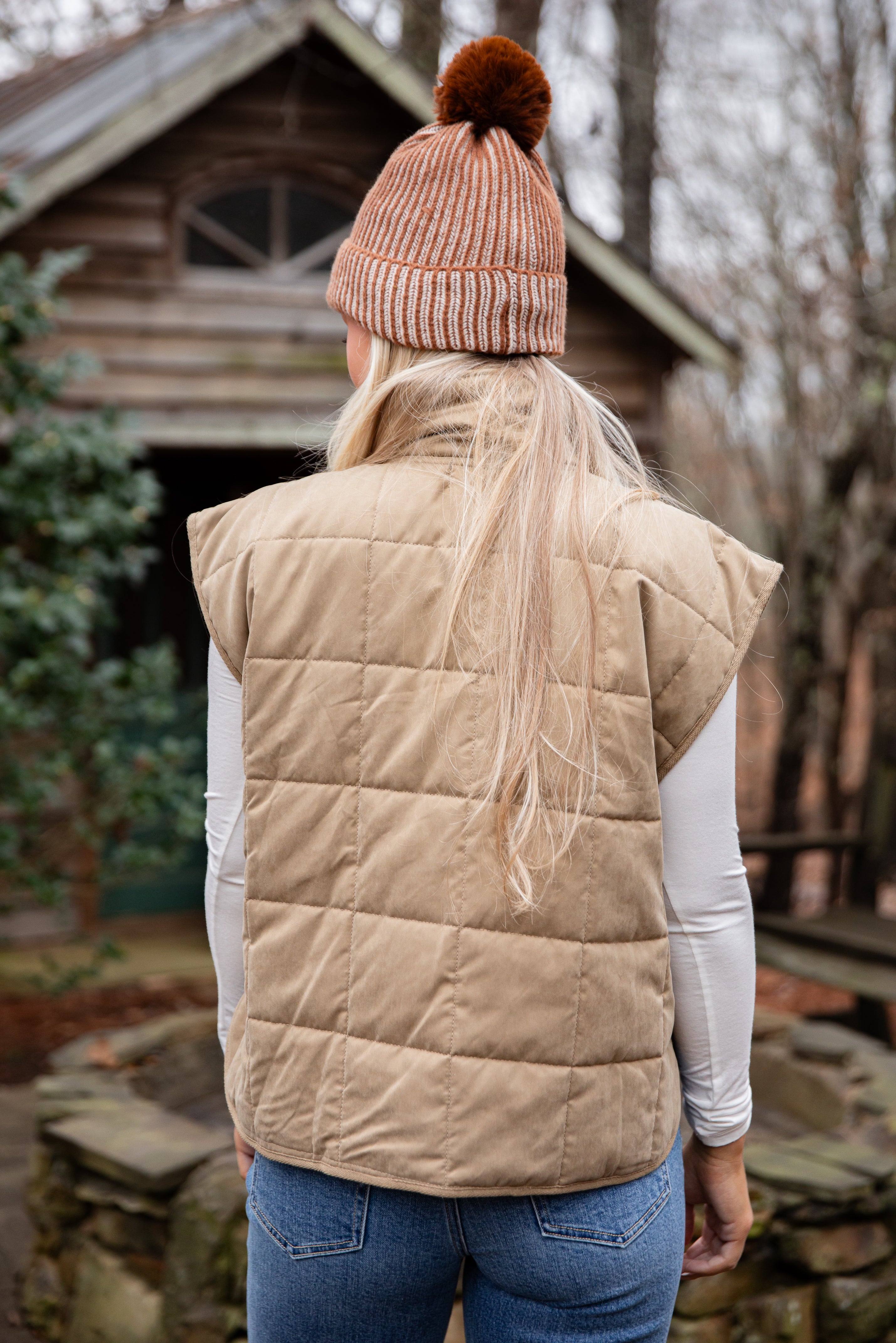 Person wearing a beige puffer vest, brown knit beanie with pom-pom, and blue jeans outdoors.