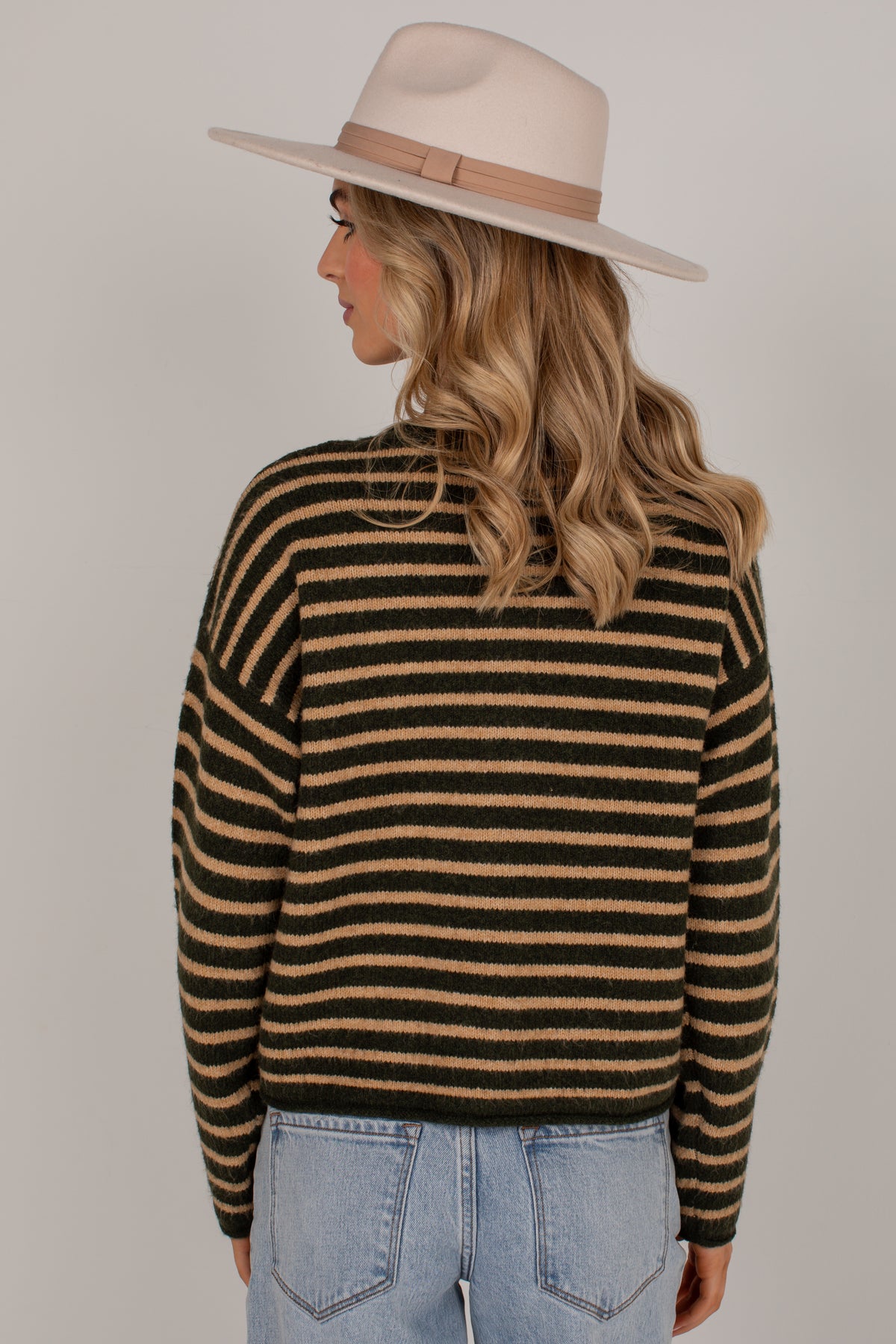 A woman shown from the back wearing a black and tan striped knit sweater, a cream wide-brim hat, and light blue jeans against a plain light gray studio background.