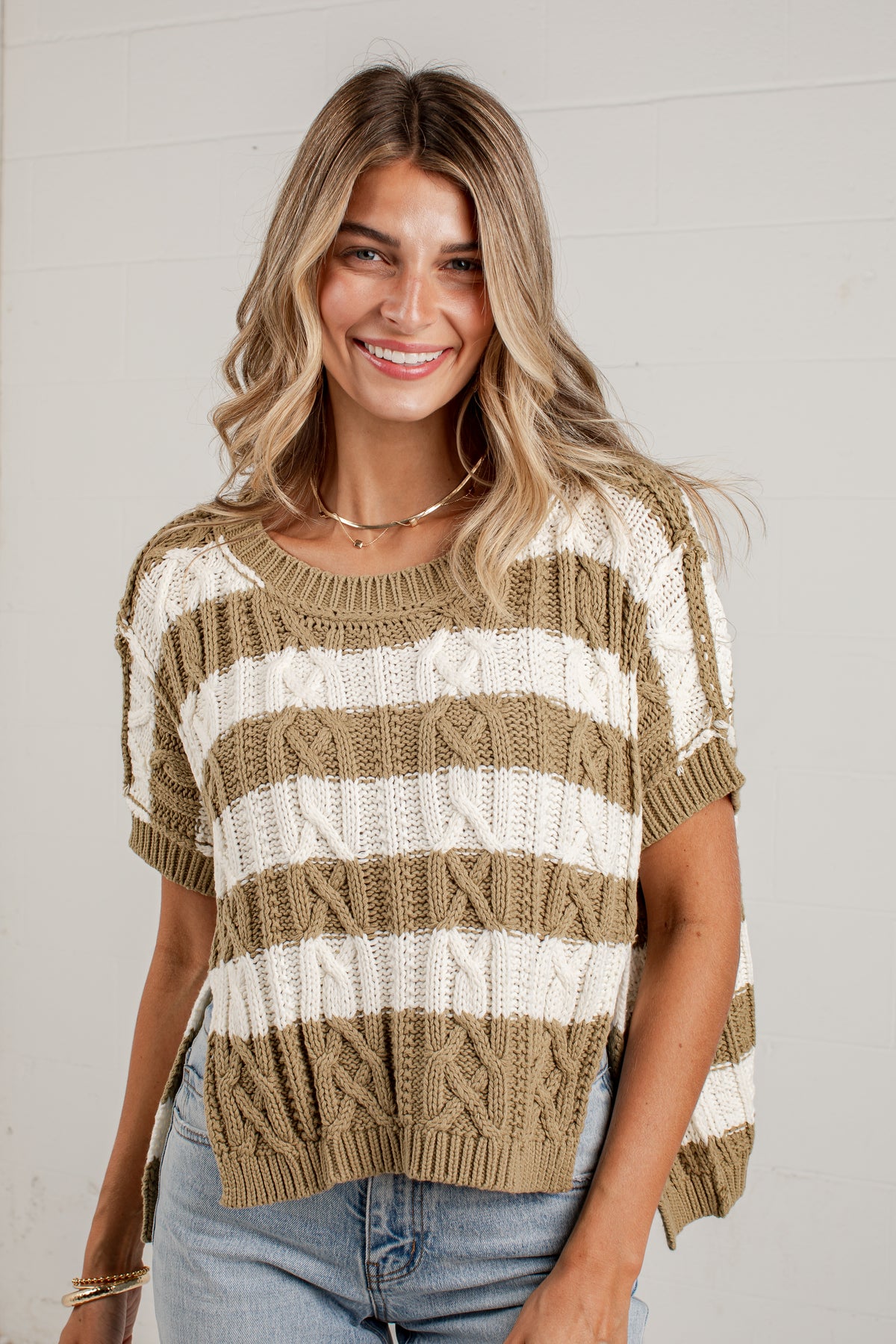 A woman wearing a short-sleeve knit sweater with horizontal beige and off-white stripes, paired with light-wash jeans, standing against a light gray wall.