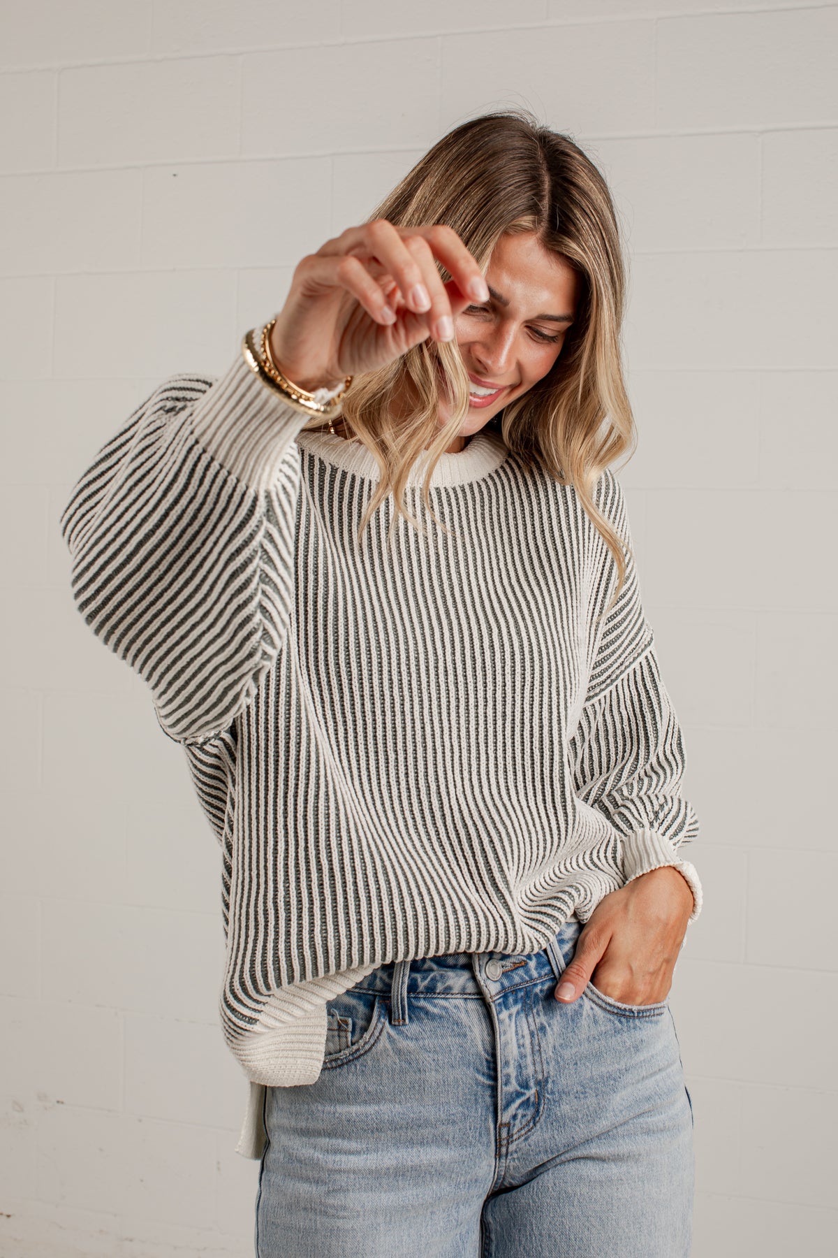 A woman wearing a cream and gray vertically striped knit sweater with a crew neck and dropped shoulders, paired with light-wash blue jeans, posing against a white paneled wall.
