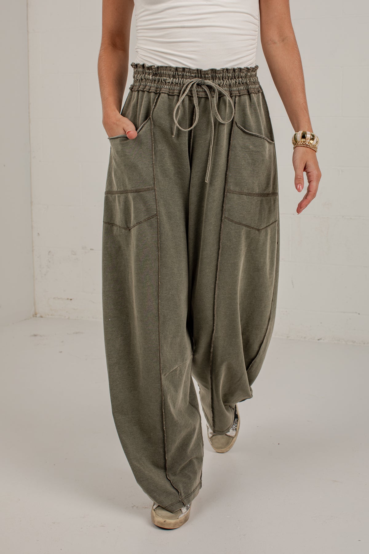 A person wearing loose, wide-leg sage green pants with an elastic drawstring waist, side pockets, and a relaxed drape, paired with a white top and beige shoes in a studio setting.
