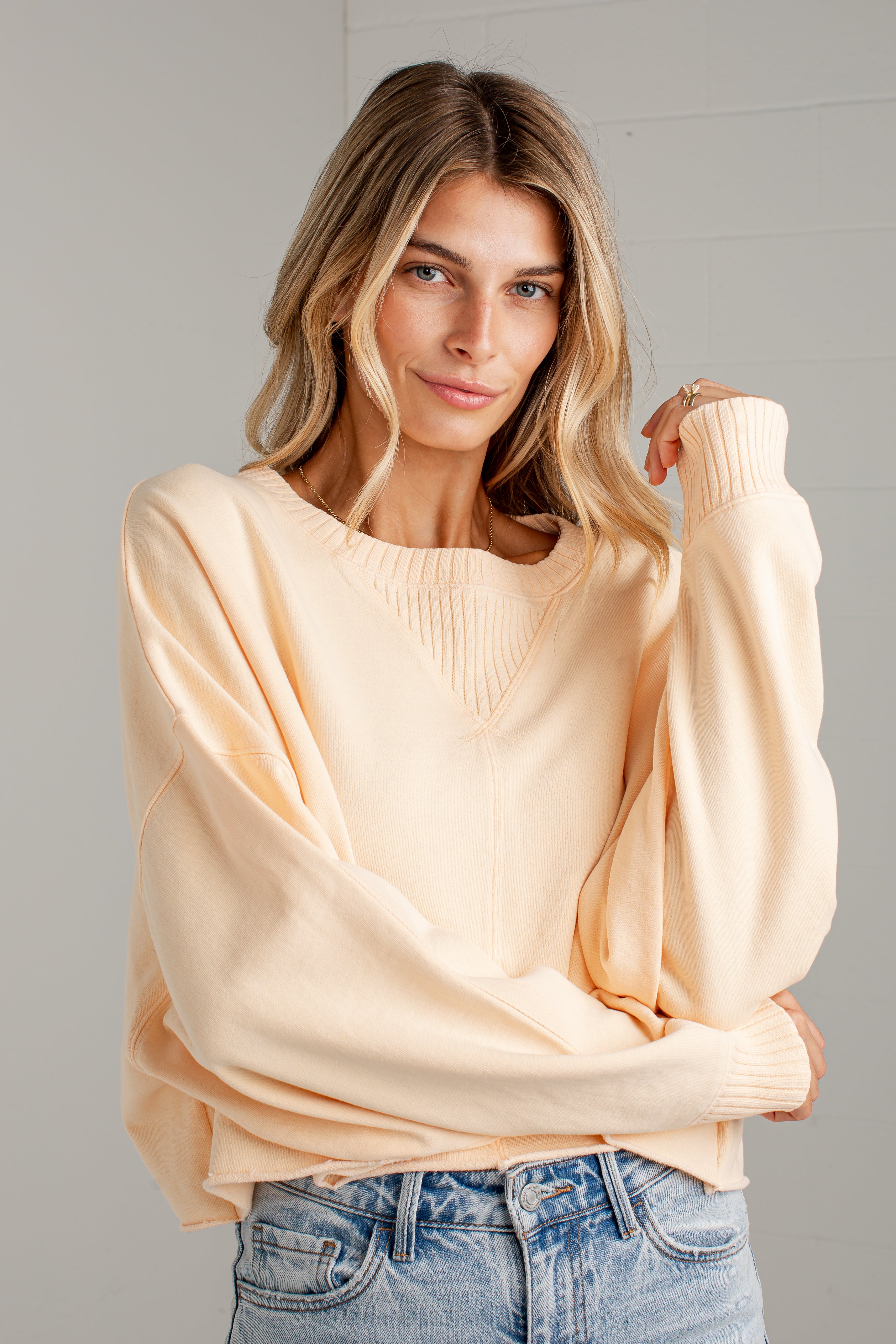 A woman wearing a light peach-colored knit sweater with balloon sleeves and a ribbed V-neck, paired with blue jeans, posing against a light gray wall.