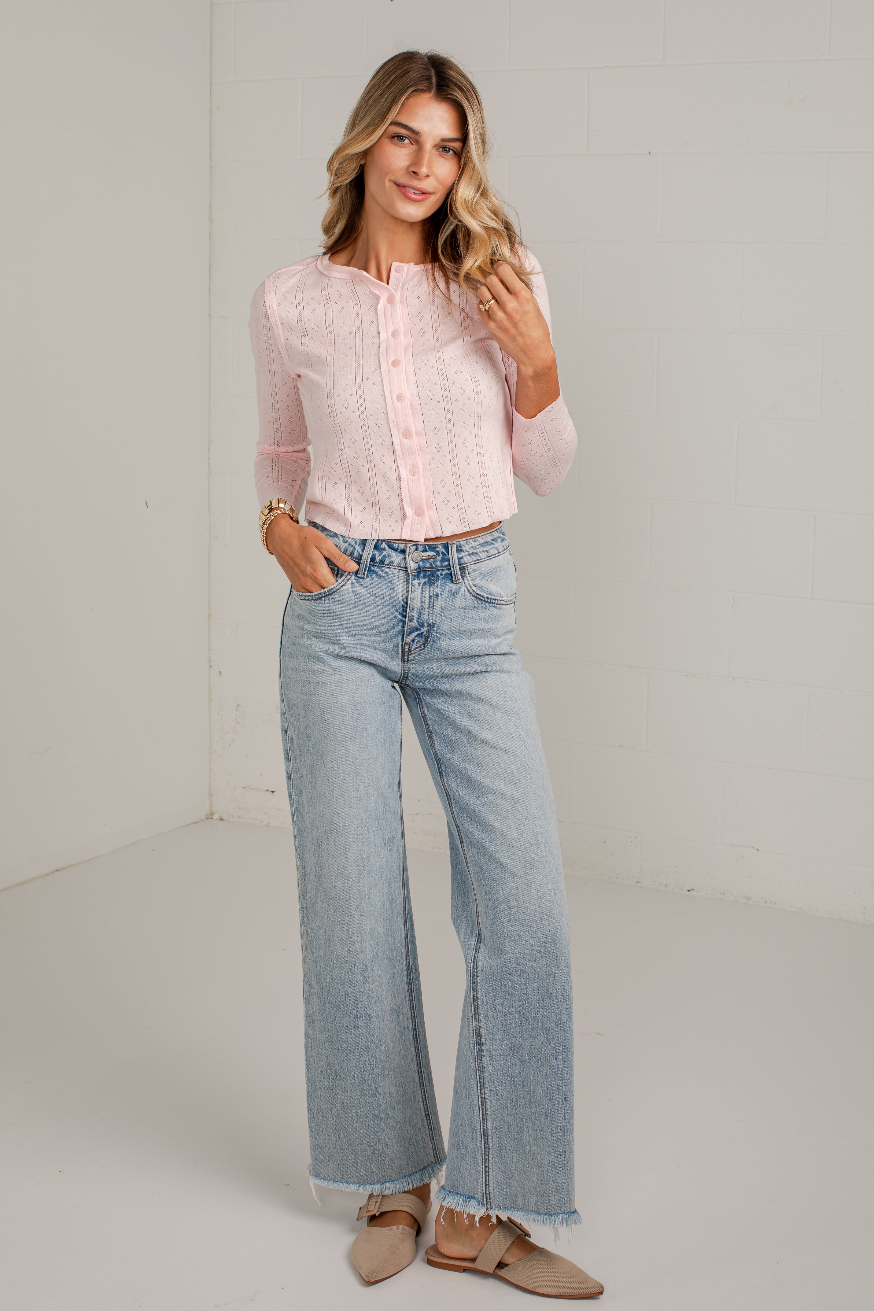 A woman modeling a light pink long-sleeve button-up blouse and light-wash wide-leg jeans, standing in a bright white studio corner.